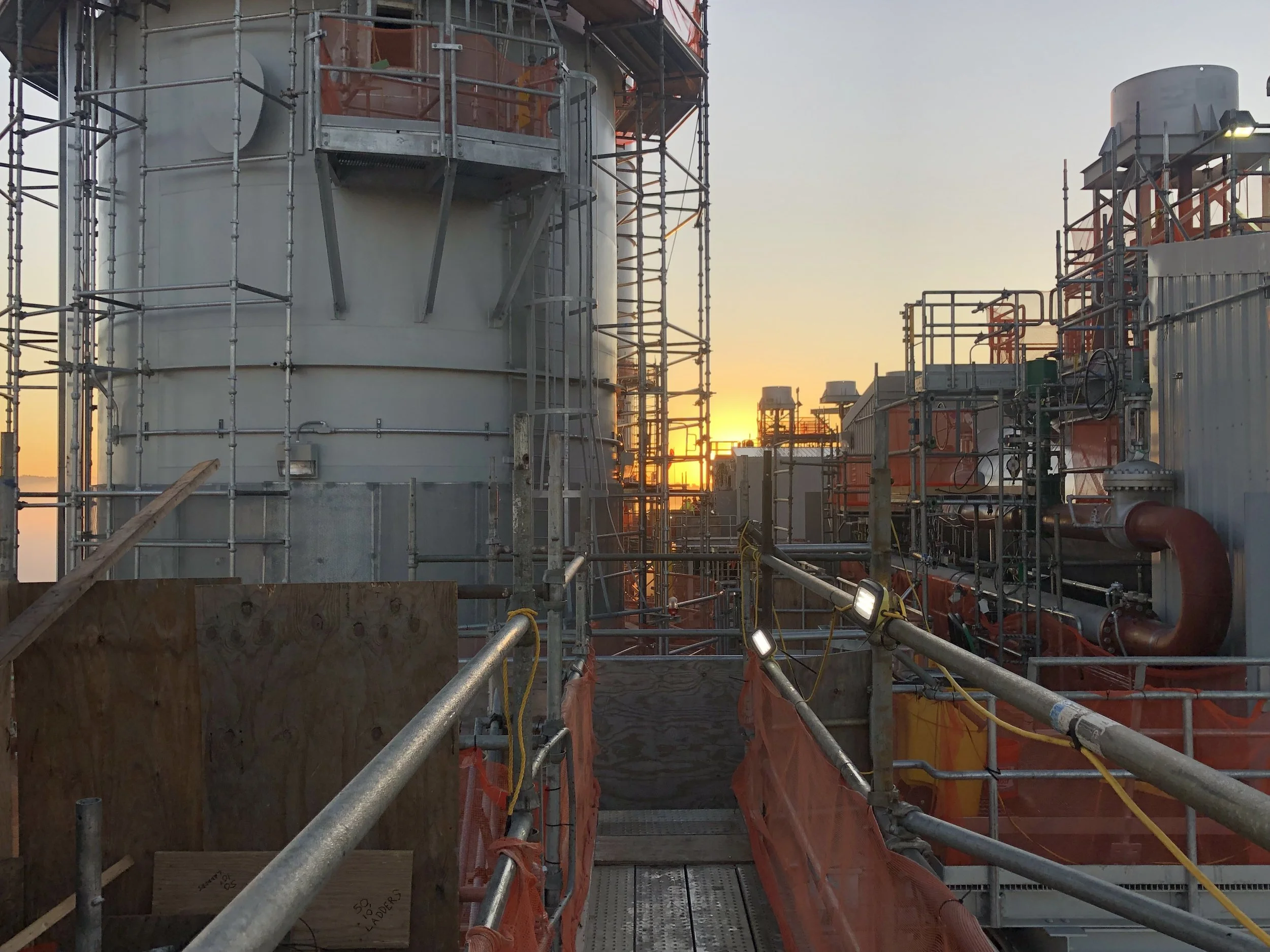 Construction site with large industrial tanks surrounded by scaffolding and piping, during sunset.