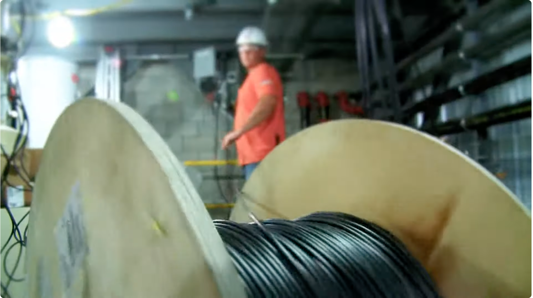 Worker in safety gear standing in an industrial setting behind large spools of wire.