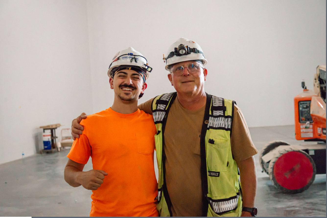 Two construction workers posing together inside a building under construction. One is a young man in an orange shirt and the other is an older man in a yellow safety vest, both wearing white hard hats with work lights, and the older man has his arm around the younger man's shoulders.