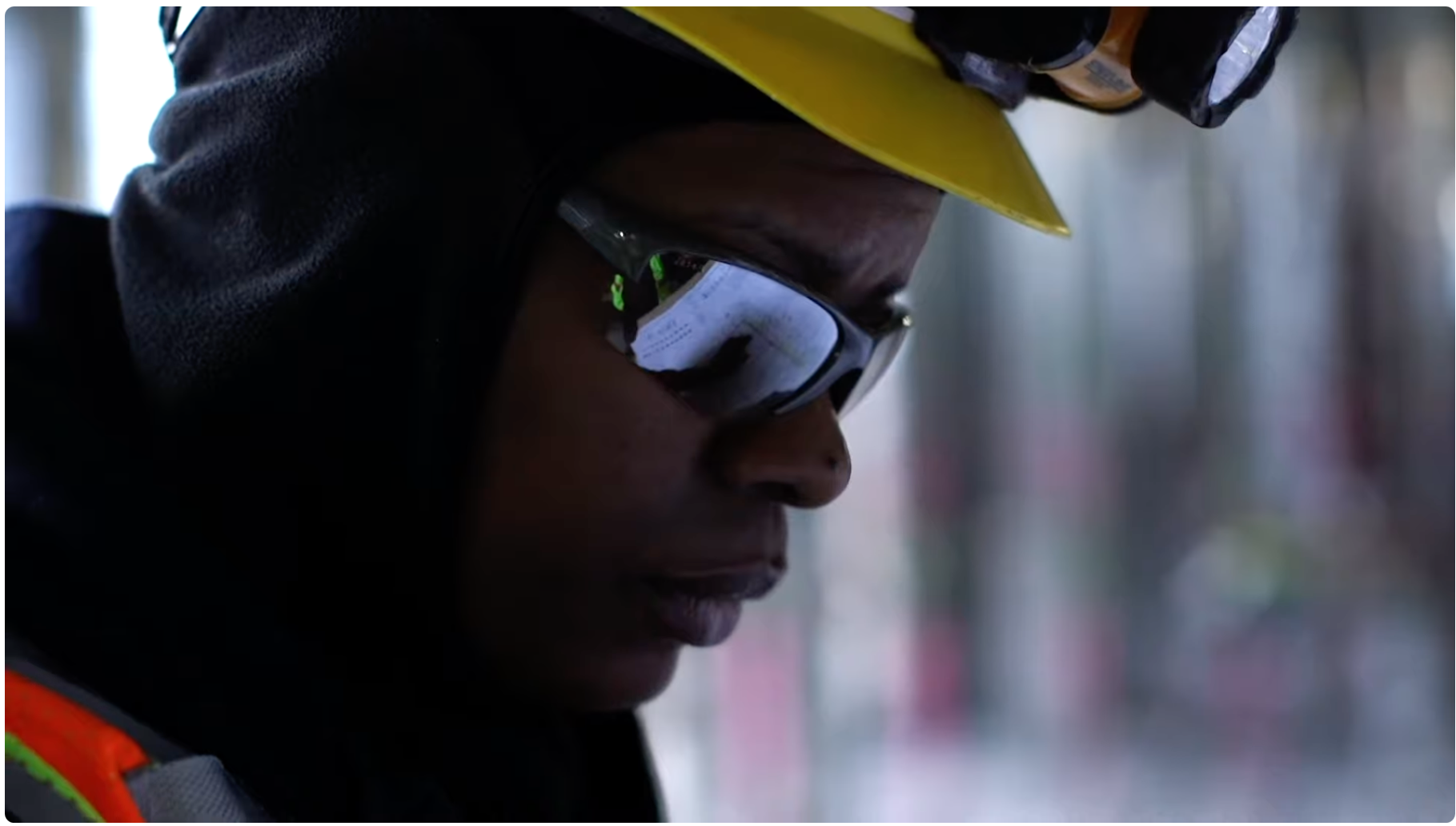 Close-up of a person wearing reflective safety glasses, a yellow hard hat, and a black hooded jacket.
