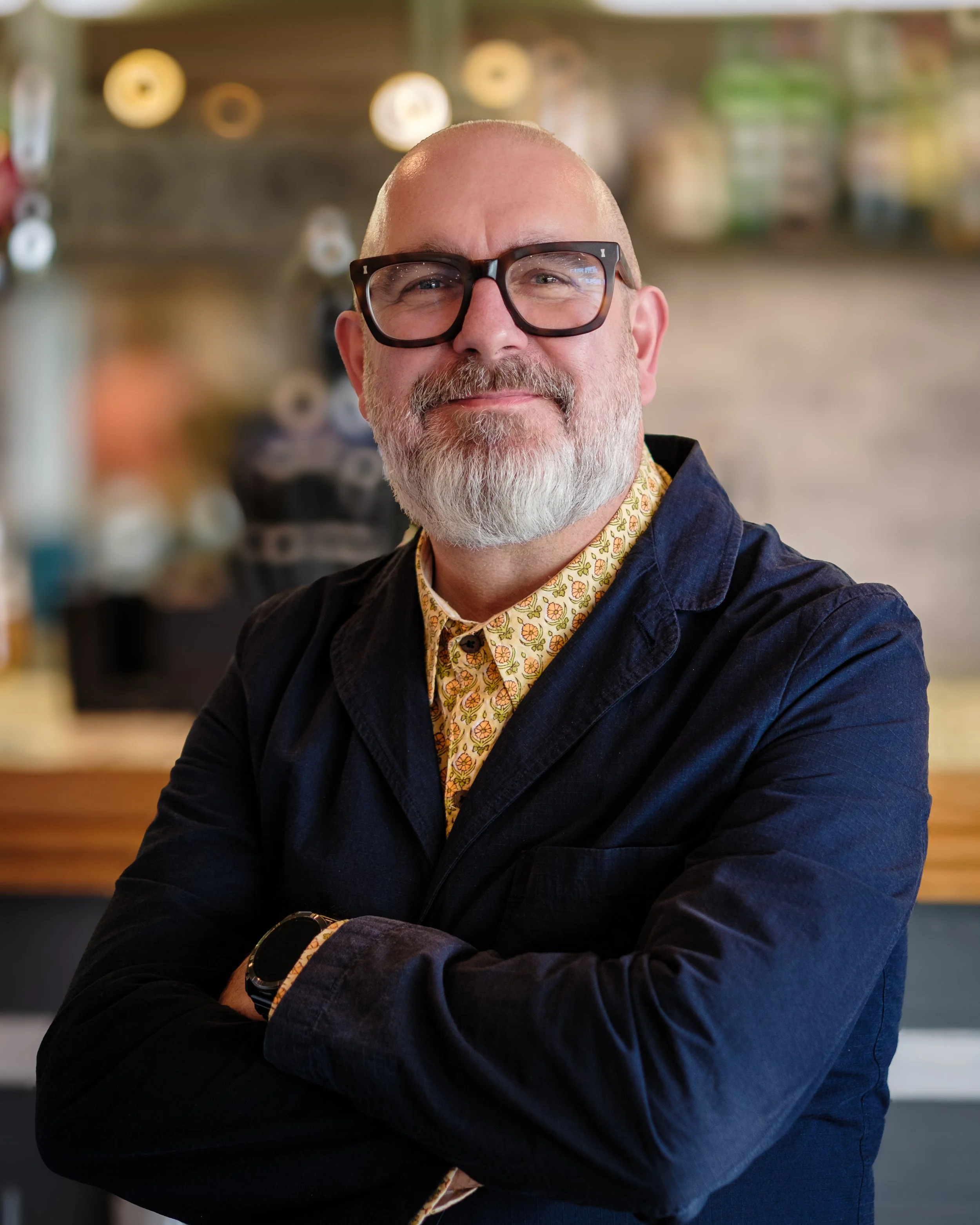 A middle-aged man with a beard and glasses, wearing a colorful shirt and dark jacket, smiling at the camera with crossed arms in a cafe or restaurant with ambient lighting.