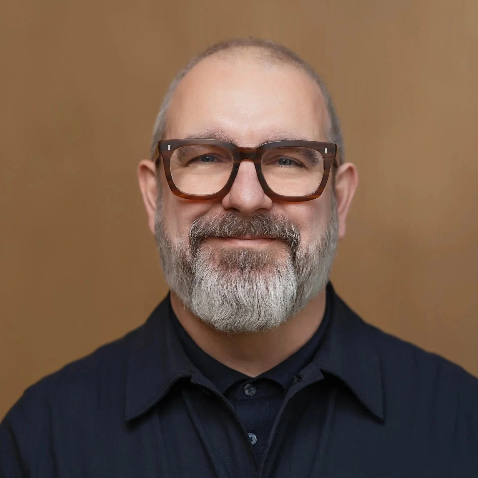 Close-up portrait of a middle-aged man with glasses, gray beard, and receding hair, wearing a black shirt, smiling softly against a plain brown background.