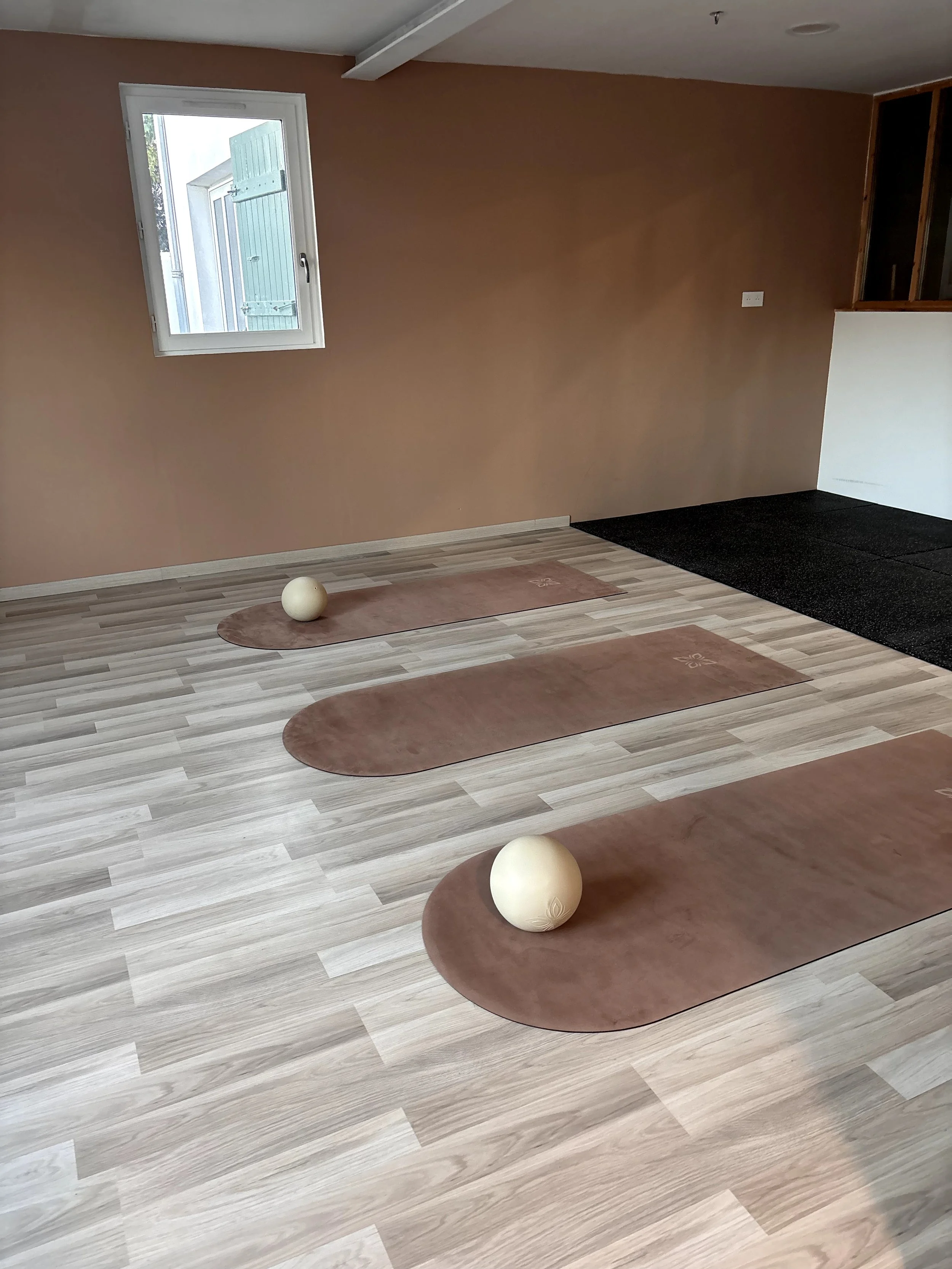 Salle de yoga ou méditation avec tapis en terre cuite, balles en bois et mur beige, fenêtre avec vue sur l'extérieur.