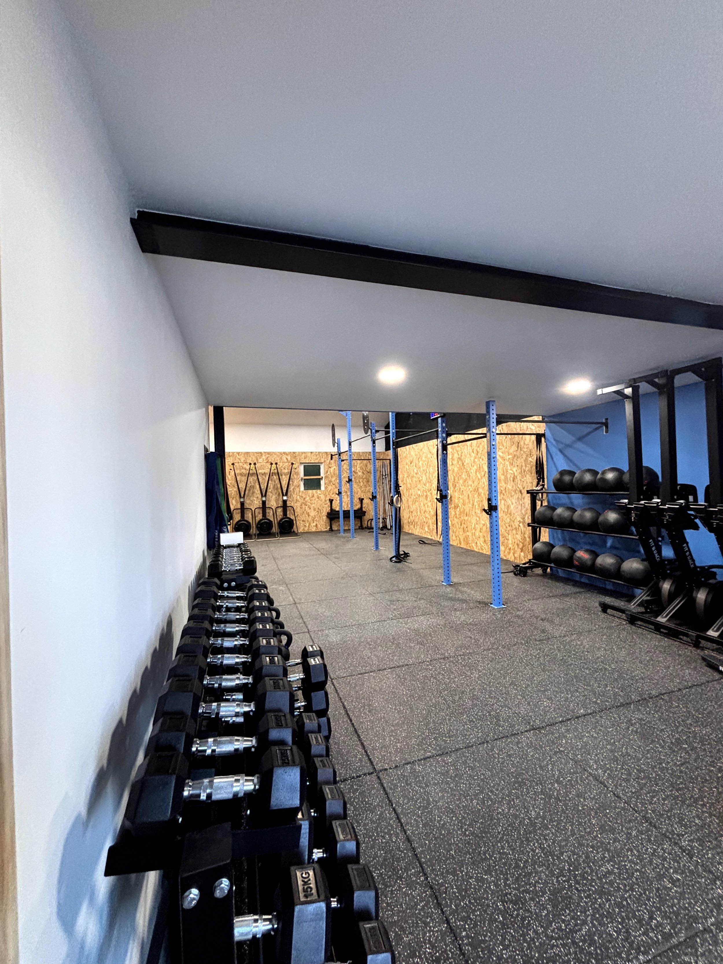 Salle de sport avec haltères, kettlebells et équipements de musculation sur un sol en caoutchouc, murs blanche et en bois, éclairage lumineux.
