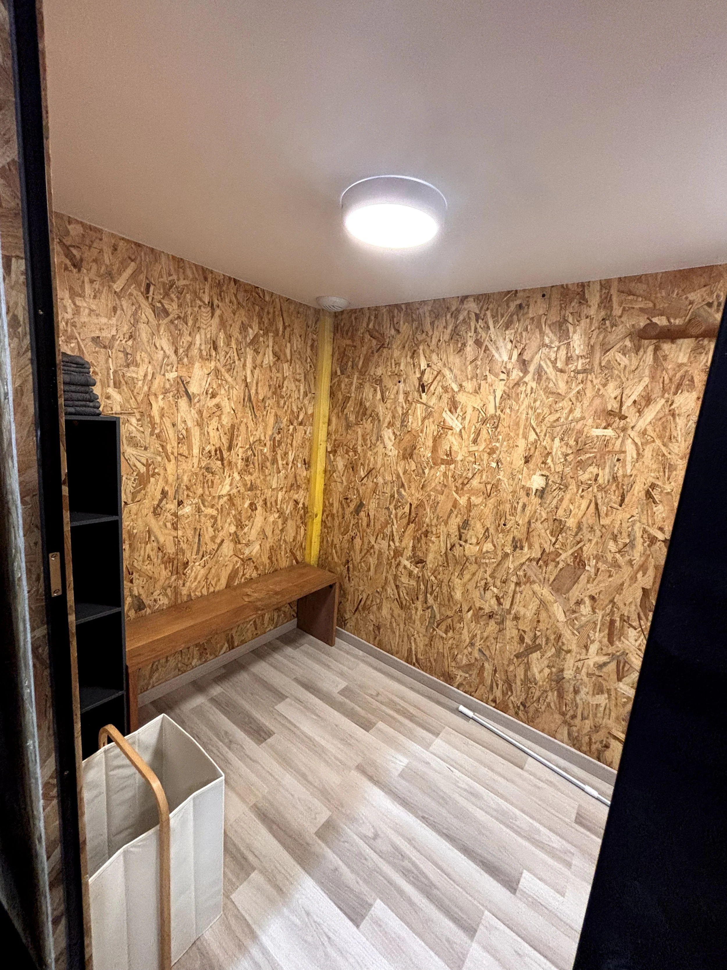 Une pièce en cours de rénovation avec un mur recouvert de panneaux en bois, un banc en bois, une étagère noire avec des serviettes, un seau blanc, et des outils au sol.