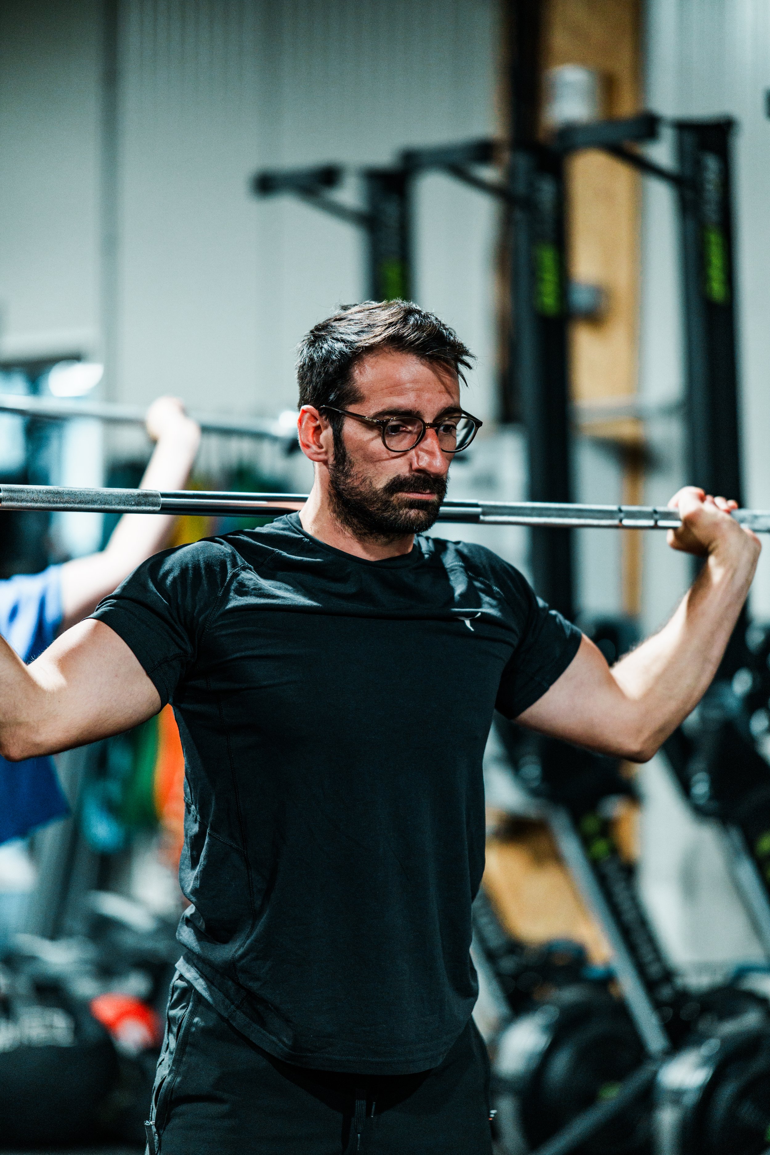 Un homme musclé, portant des lunettes et un t-shirt noir, fait un entraînement de musculation avec une barre dans une salle de sport. Il semble concentré et déterminé pendant sa séance d'exercice.