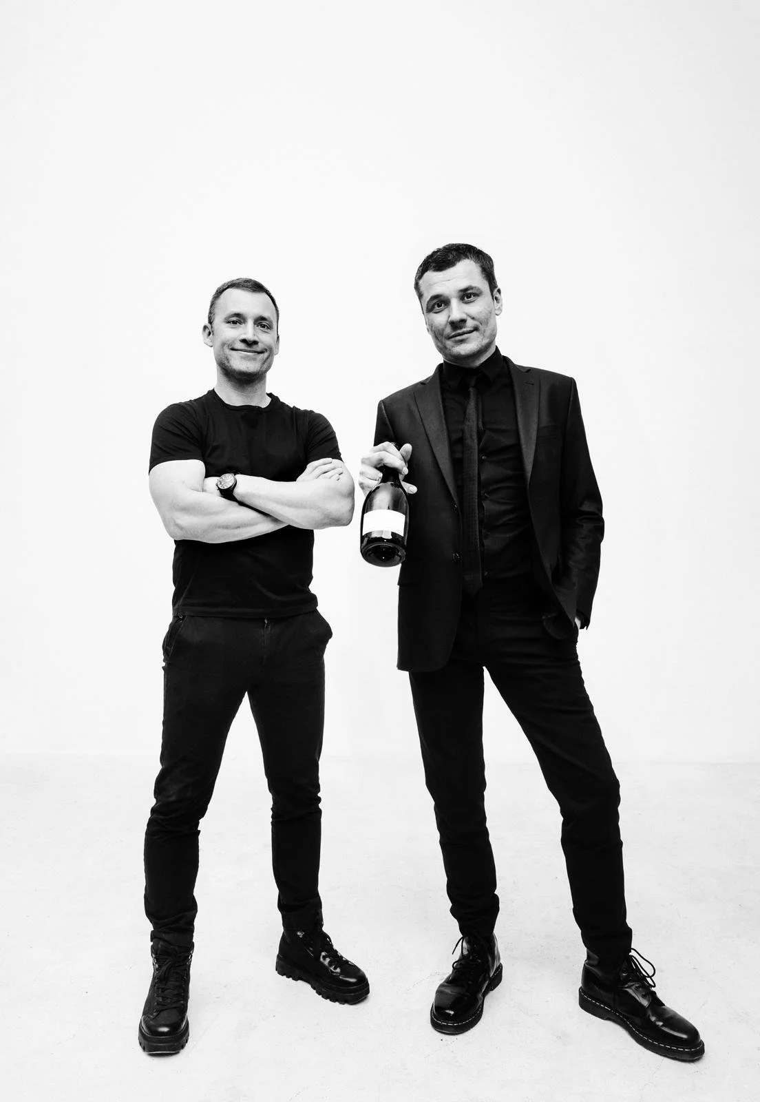 Two men standing against a plain white wall, one with arms crossed in casual attire, the other holding a wine bottle in a suit.