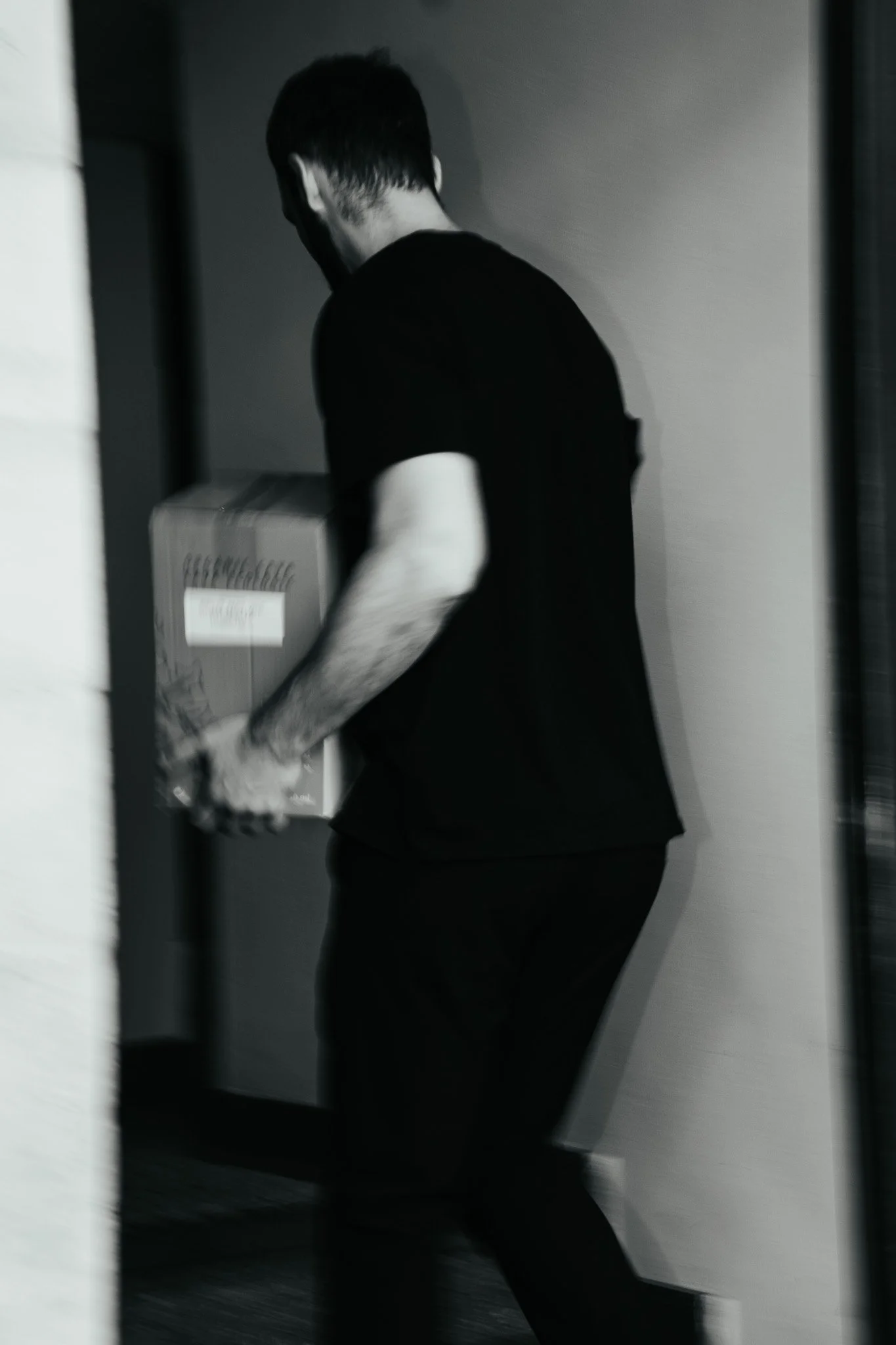 A person with black hair, wearing a black shirt, carrying a cardboard box while walking through a doorway.
