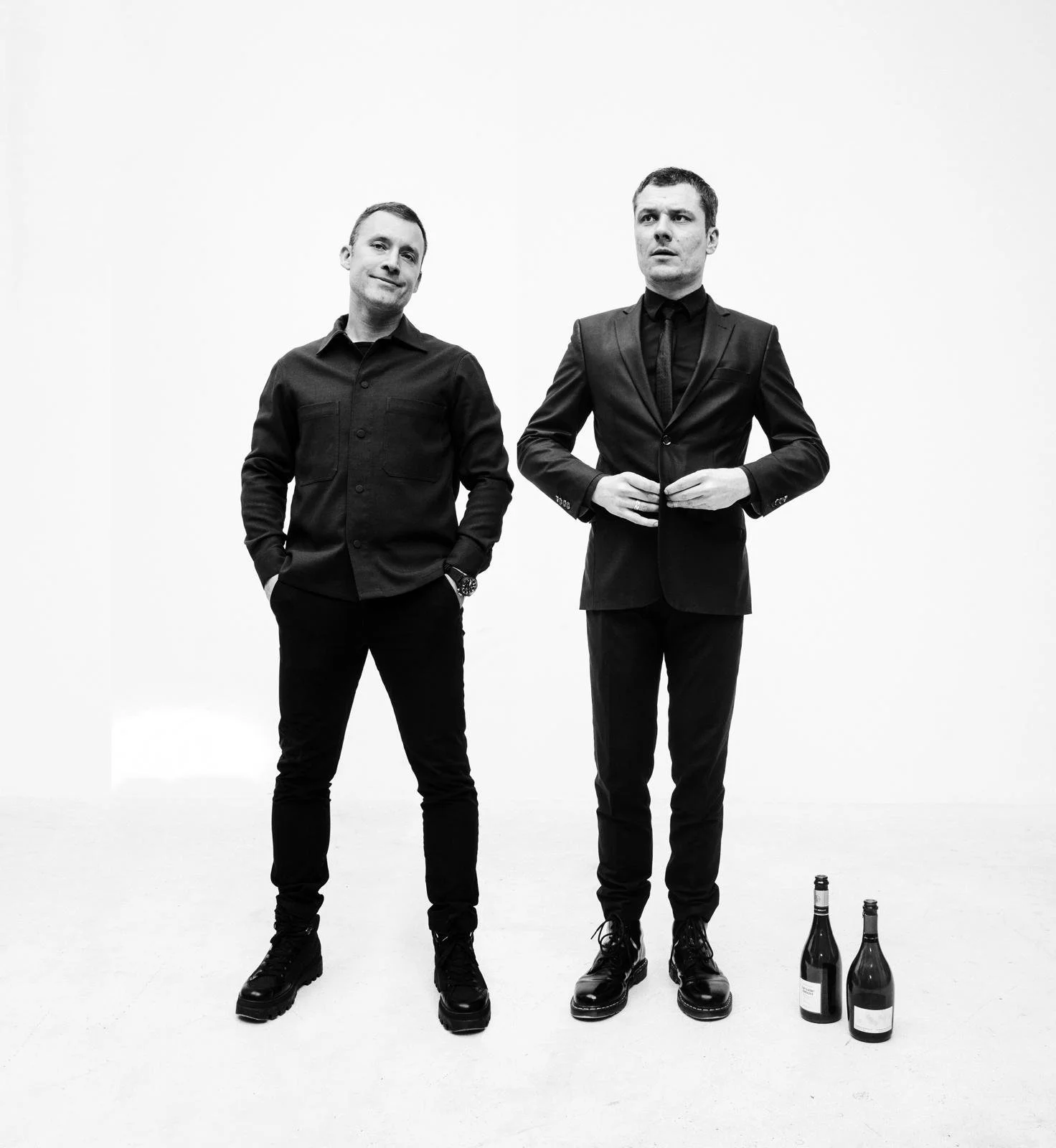 Two men in black clothing and boots standing in front of a plain white background, with two bottles of wine placed on the floor near them.