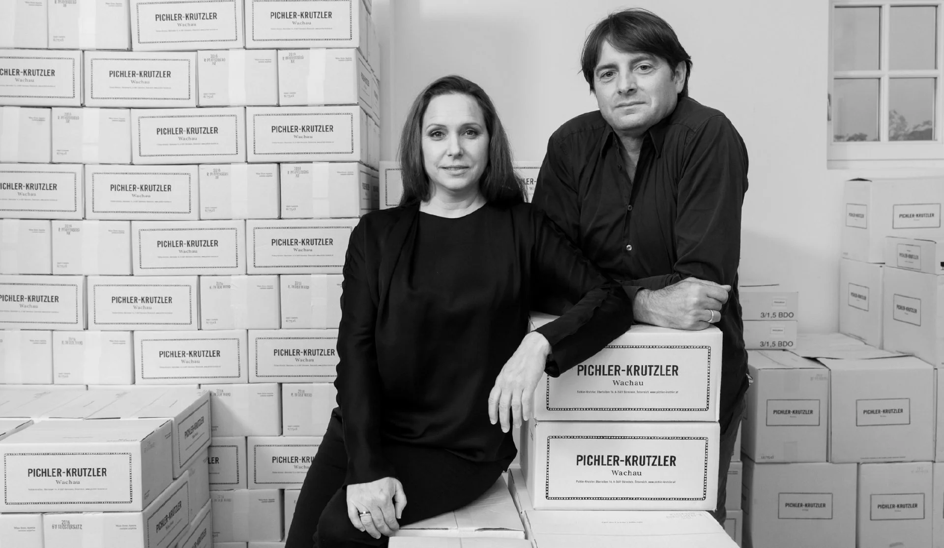 A black and white photo of a woman and a man posing in a room filled with stacked cardboard boxes labeled 'PICHLER-KRUTZLER' and 'Wachau'. The woman has shoulder-length dark hair and is wearing a dark top, standing with her hand resting on a box. The man has dark hair parted to the side, wearing a dark shirt, and is leaning on a box with his arms crossed. A window is visible in the background.