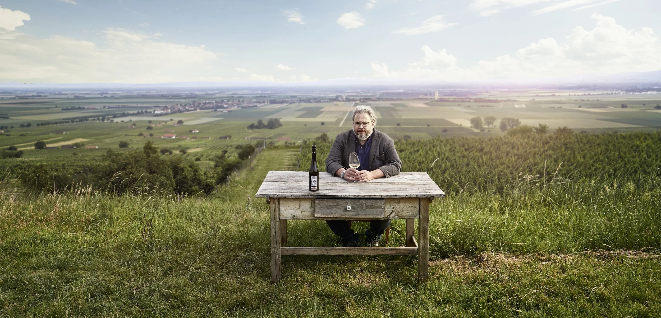 A man sitting at a rustic wooden table in a vineyard, holding a glass of white wine, with a bottle of wine on the table, overlooking lush green rolling fields and distant town under a partly cloudy sky.