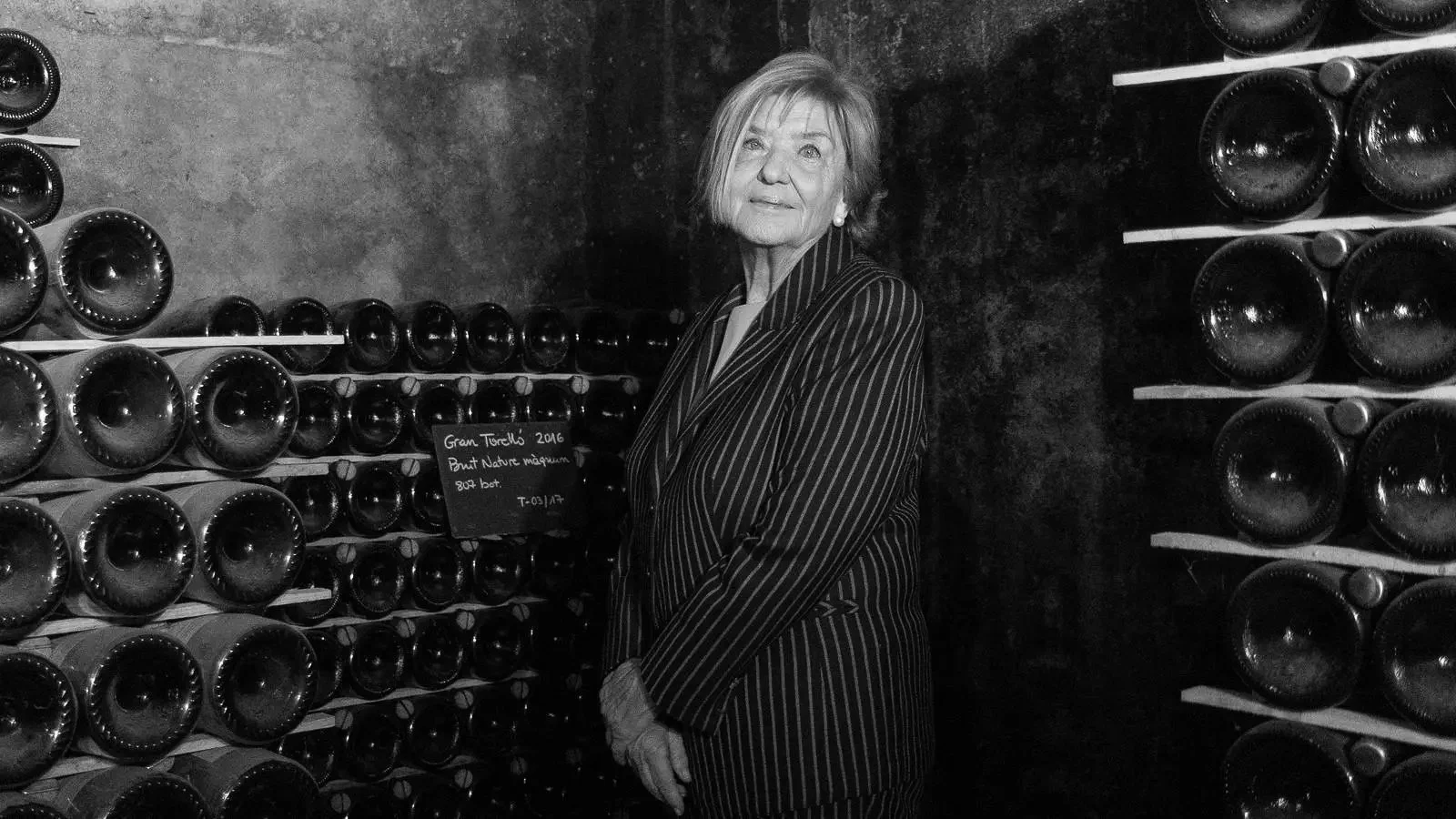 An elderly woman in a pinstripe blazer standing among wine bottles in a wine cellar, looking up with a slight smile.