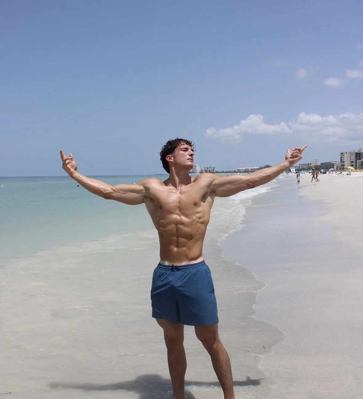 A shirtless man with muscular build and blue shorts standing on a sandy beach with arms outstretched, facing the ocean with a clear blue sky in the background.