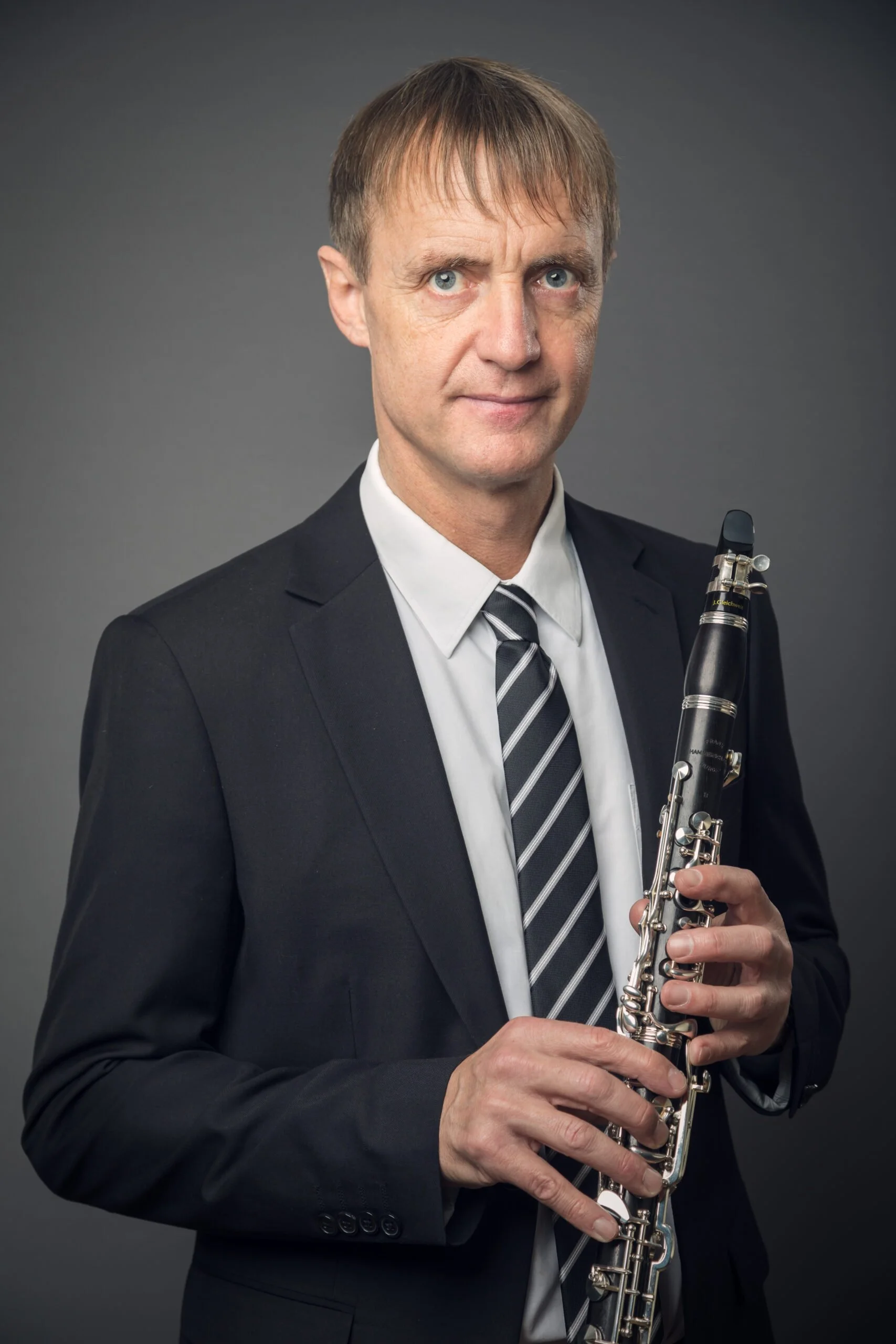A middle-aged man in a dark suit holding a clarinet against a plain gray background.