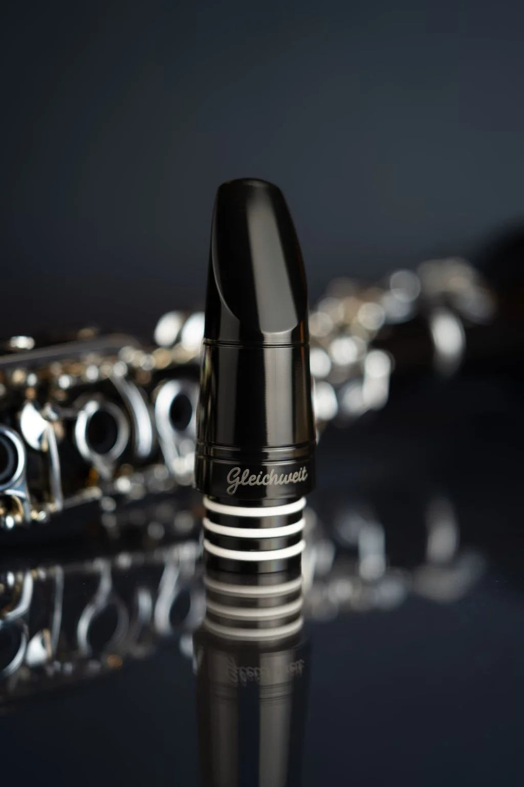 Close-up of a black musical instrument mouthpiece with the word 'Gleichweit' written on it, positioned in front of a blurred shiny flute.