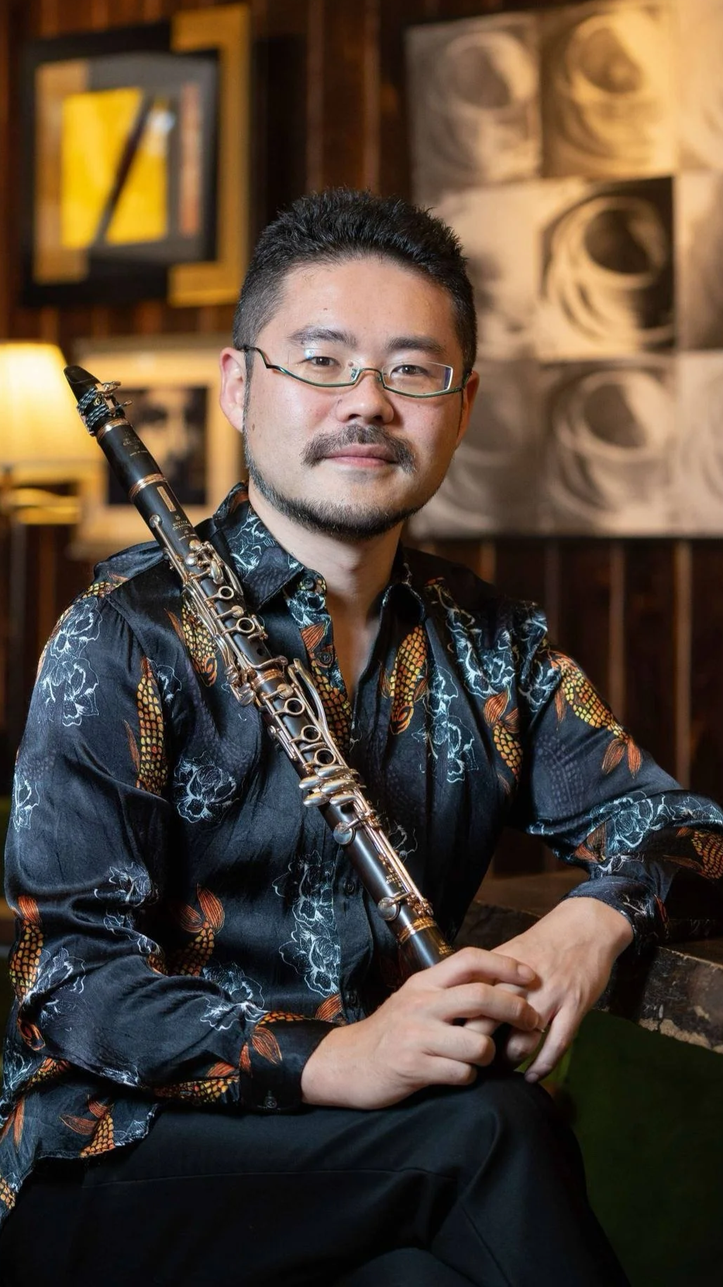 Man with glasses holding a clarinet, sitting in a room with framed artwork and warm lighting.