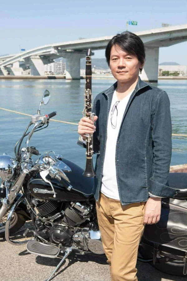 A man holding a clarinet standing next to a motorcycle near a body of water, with a large bridge and cityscape in the background on a bright day.