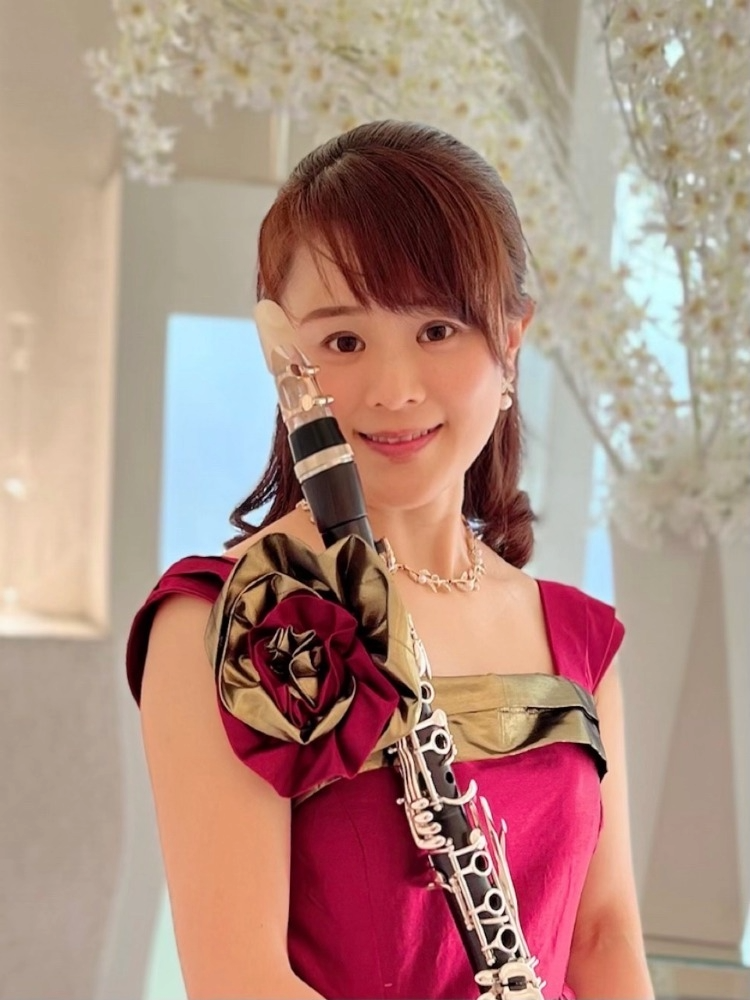 A woman in a red dress holding a clarinet, smiling indoors with white floral decorations in the background.