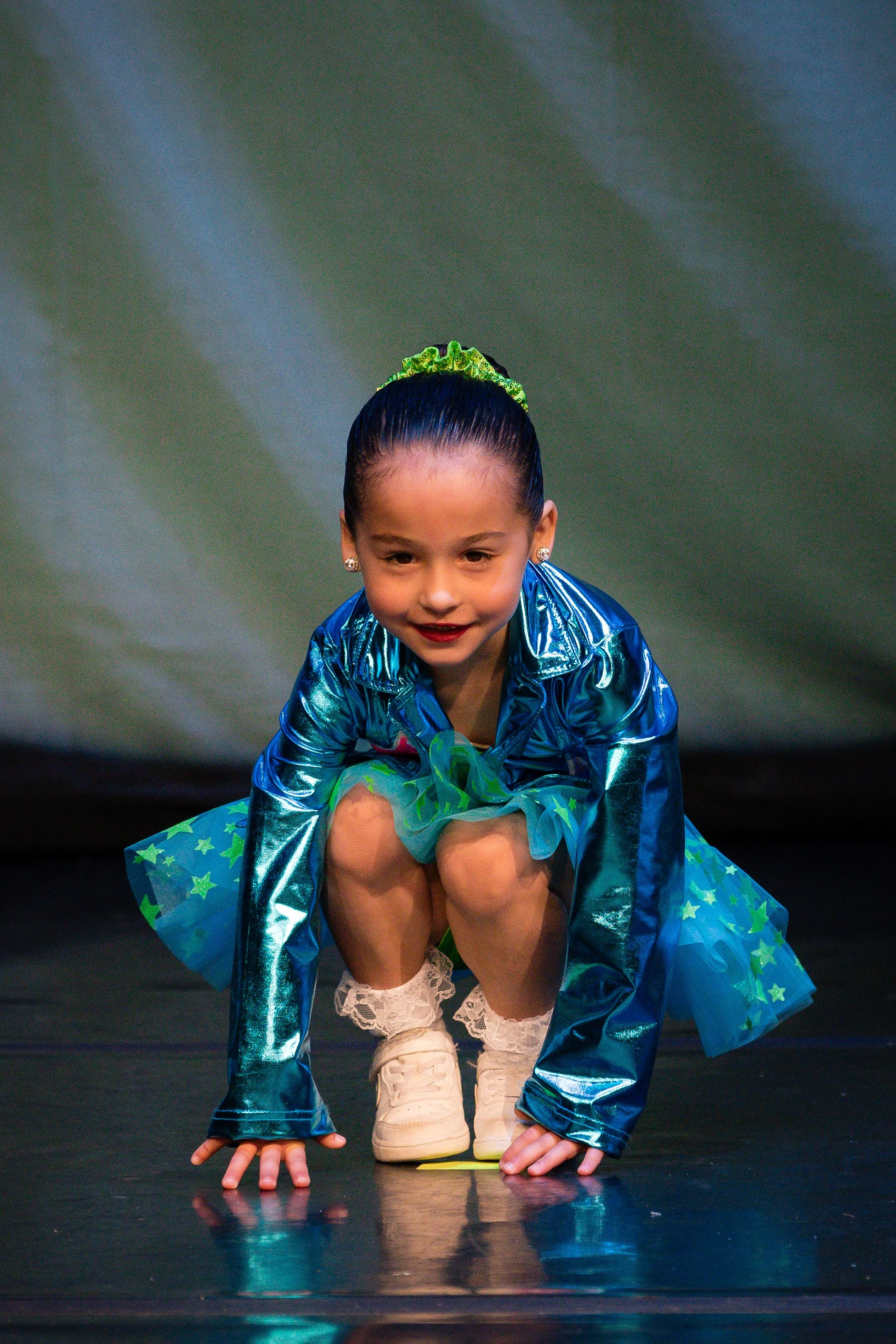 Young girl in sparkly blue costume with starry skirt performing on stage, crouched down with hands on the floor, smiling and looking forward.