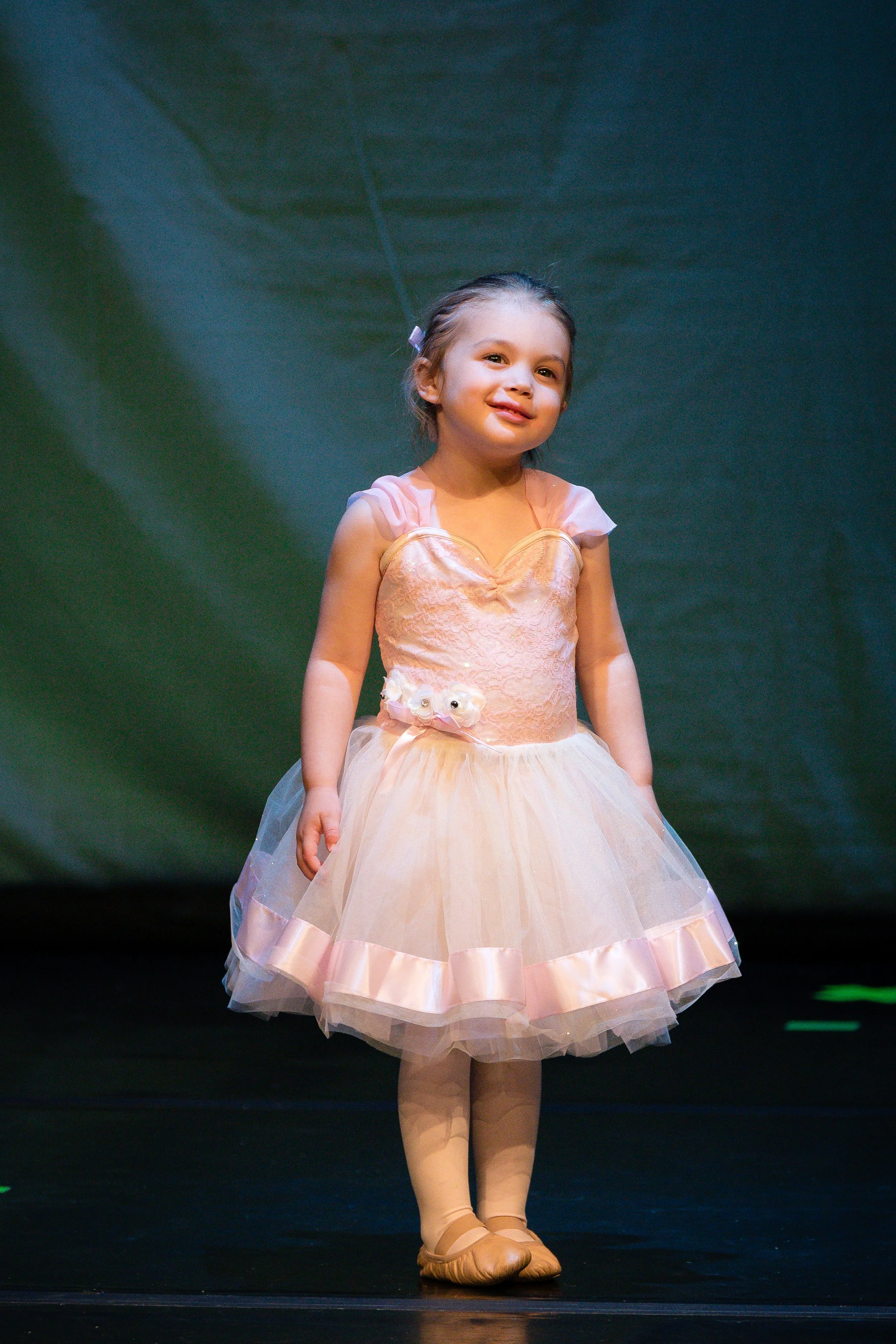 A young girl dressed as a ballerina in a peach tutu and ballet shoes standing on stage.