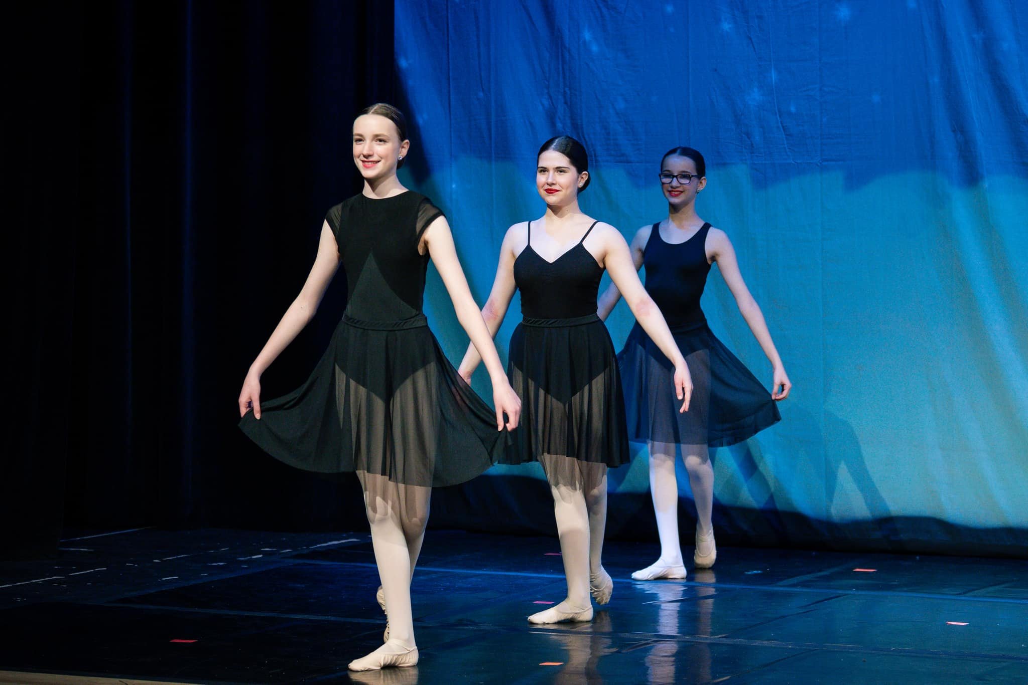 Three ballerinas in black dresses performing on stage with blue backdrop, holding hands and smiling.