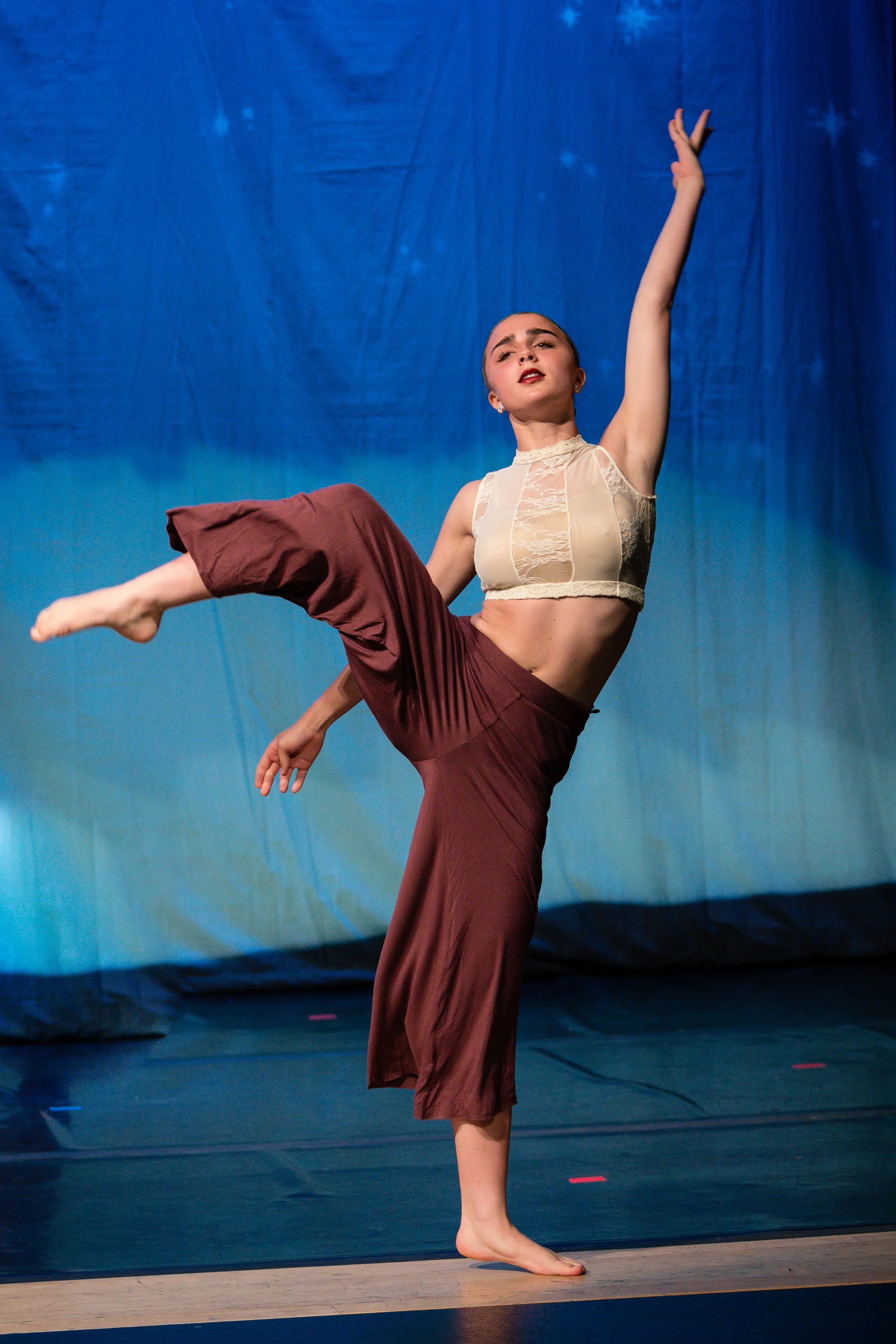 A female dancer performing a pose on stage with a blue curtain backdrop, wearing a sheer beige top and brown pants, standing on one foot with her other leg extended and raised, and one arm reaching upward.