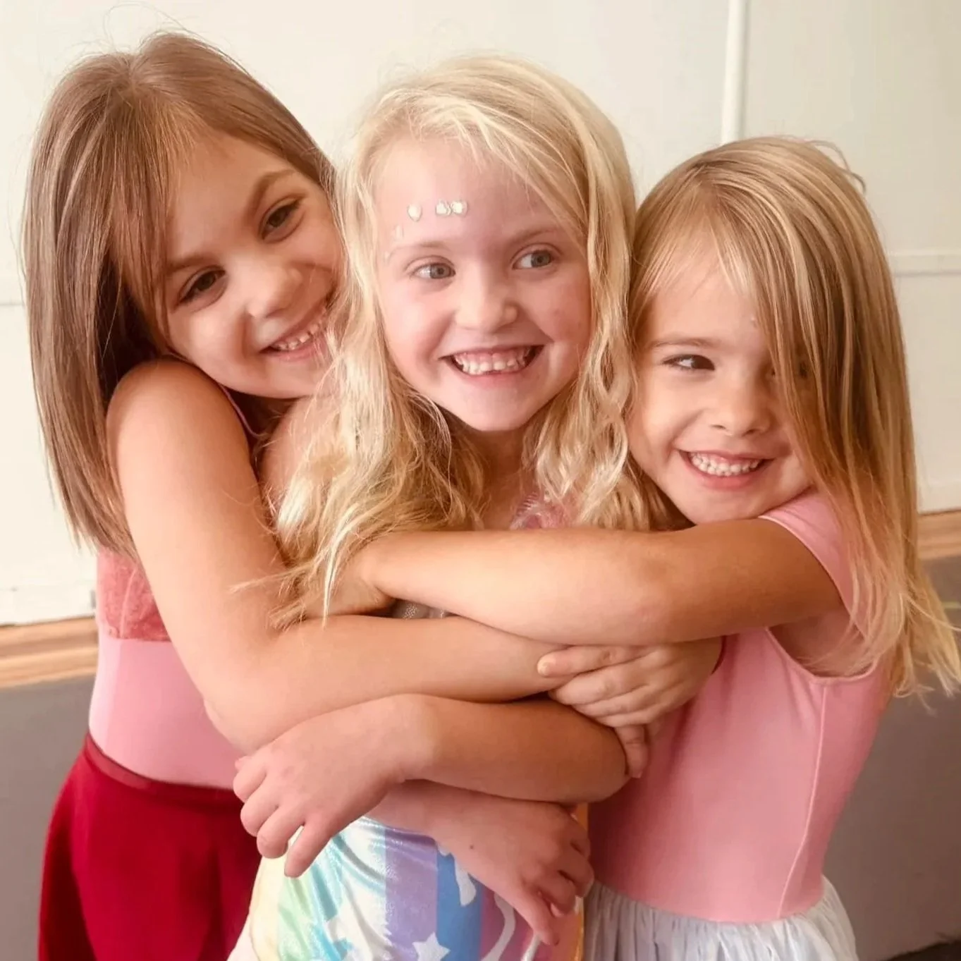 Three young girls hugging each other and smiling beaming in a close-up indoor setting.