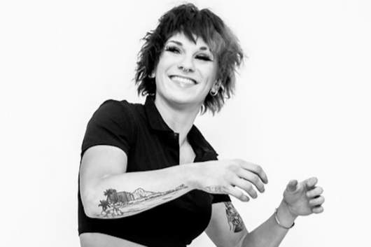 Black-and-white photo of a smiling woman with short, curly hair and tattoos on her arms, wearing a black shirt.