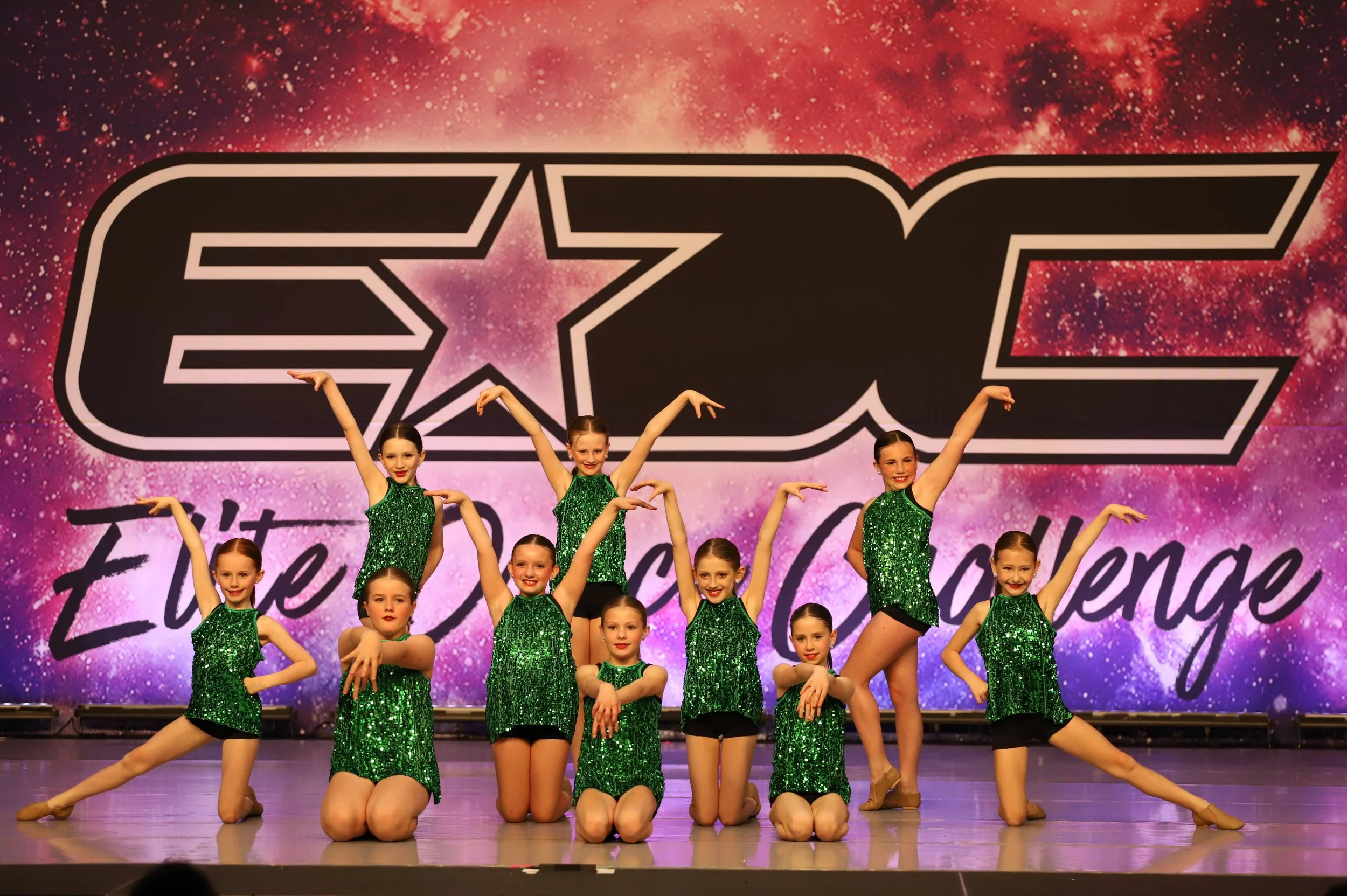 A group of young girls performing a dance on stage, wearing matching green sequined tops and black shorts, with a large backdrop displaying 'EYE' and 'Elite Dance Challenge' in the background.