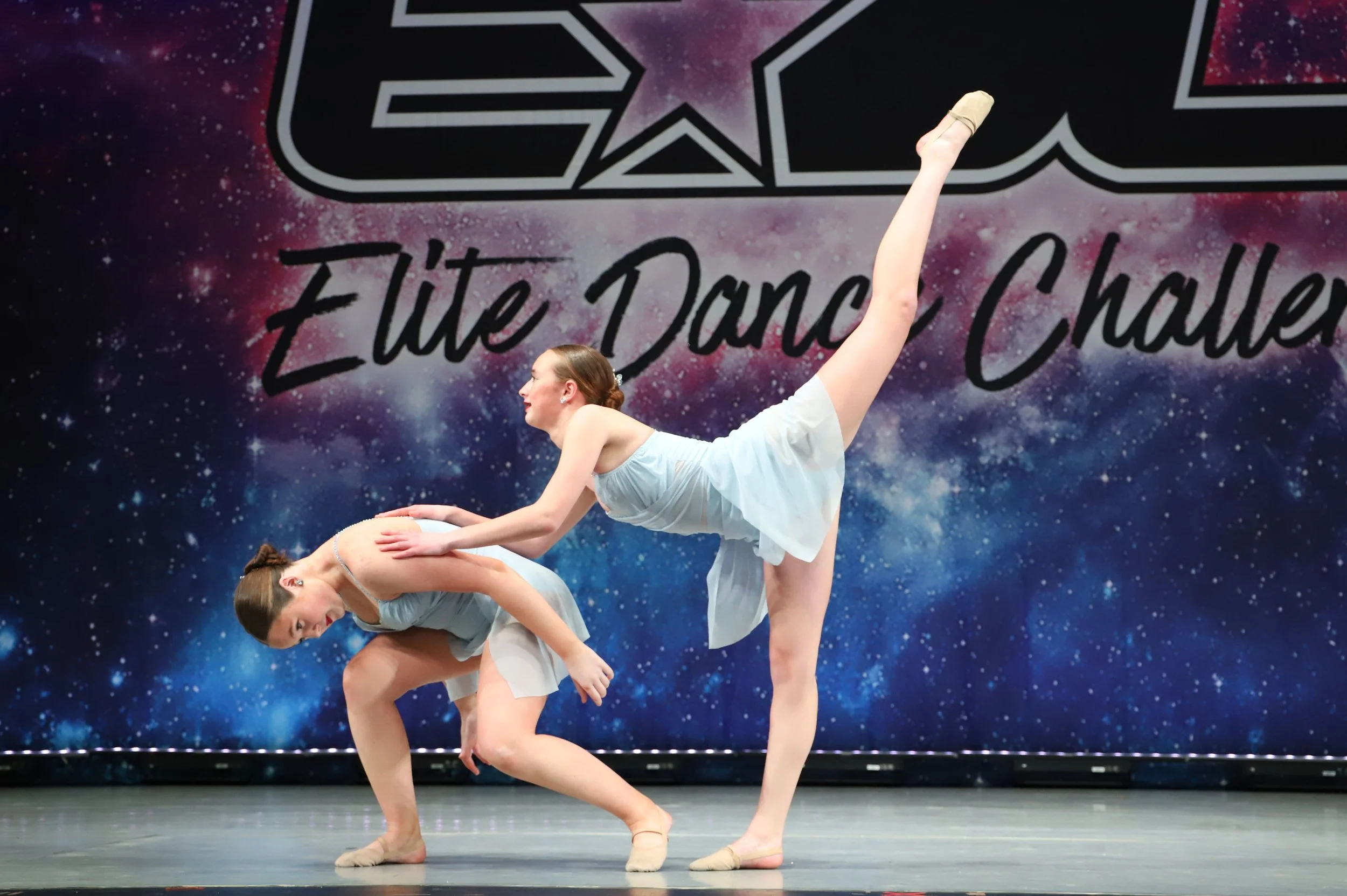 Two young female ballet dancers performing a duet on stage with a galaxy-themed backdrop that reads 'Elite Dance Challenge'. One dancer is standing on one leg with her other leg extended upwards, while the other dancer supports her by bending over an