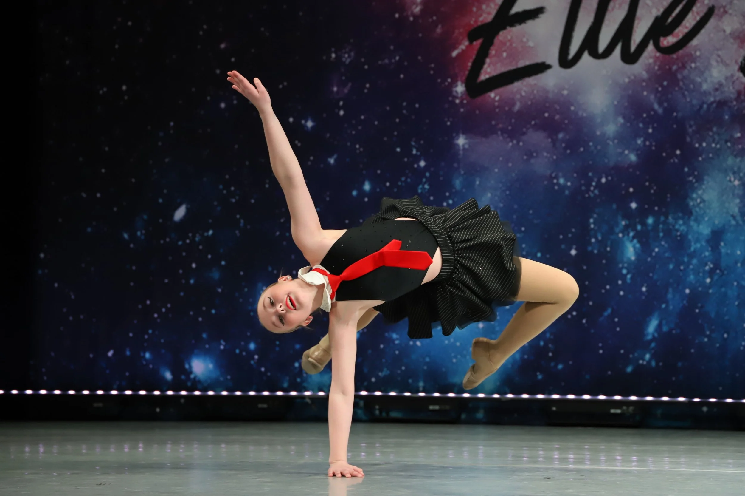 A young girl performing a dance move on stage, balancing on her right hand with her legs bent to the side, wearing a dark costume with a red scarf, against a backdrop depicting a starry galaxy with the word 'Star' written in black.