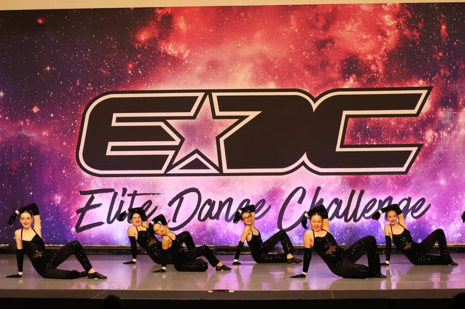 Six young female dancers in black costumes perform on stage in front of a large colorful backdrop with the words "ECE Elite Dance Challenge." The backdrop features a galaxy pattern with pink, purple, and orange hues.