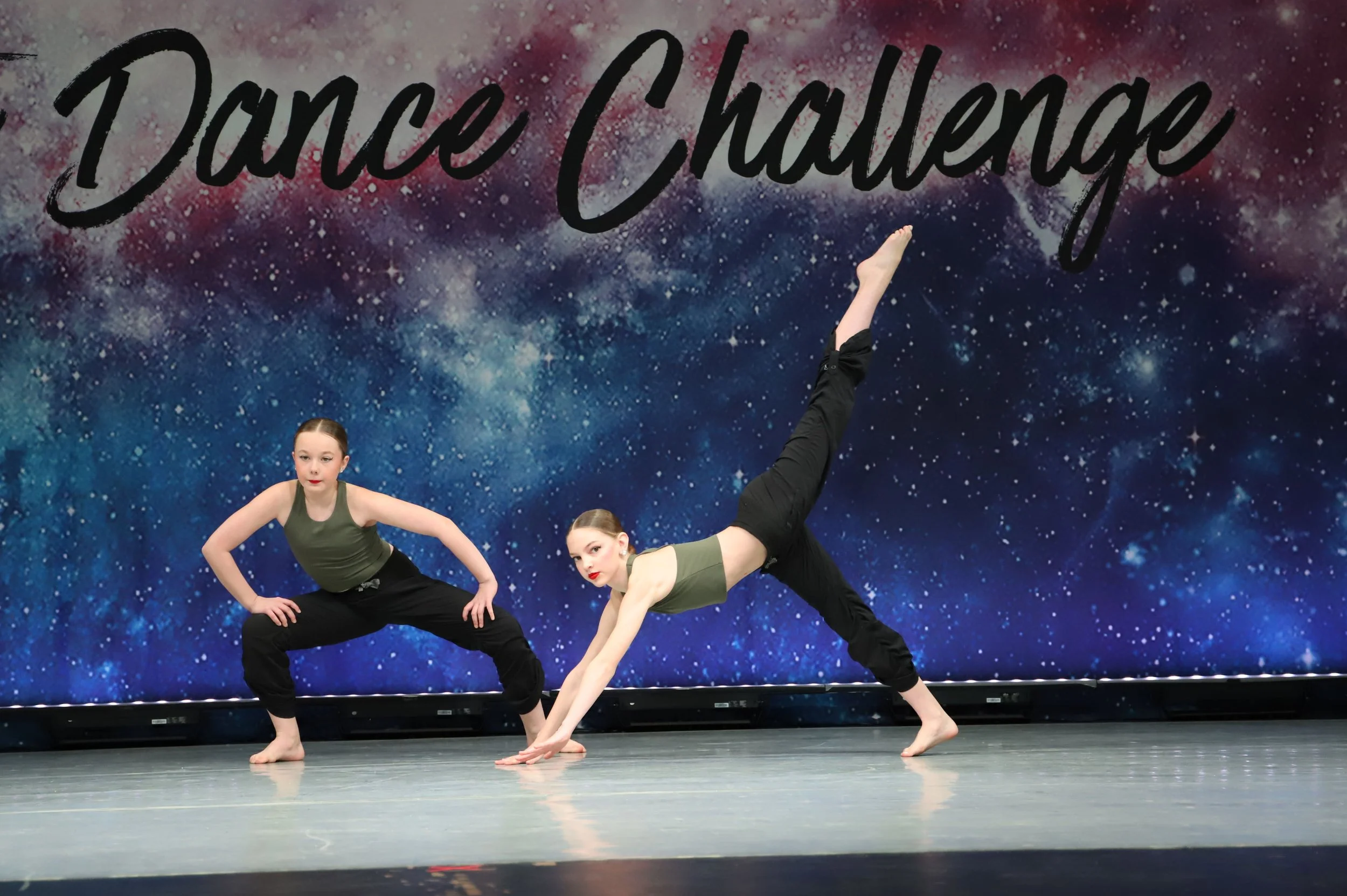Two young girls performing a contorted dance pose in front of a backdrop with a galaxy pattern and the words 'Dance Challenge' in large black cursive script.