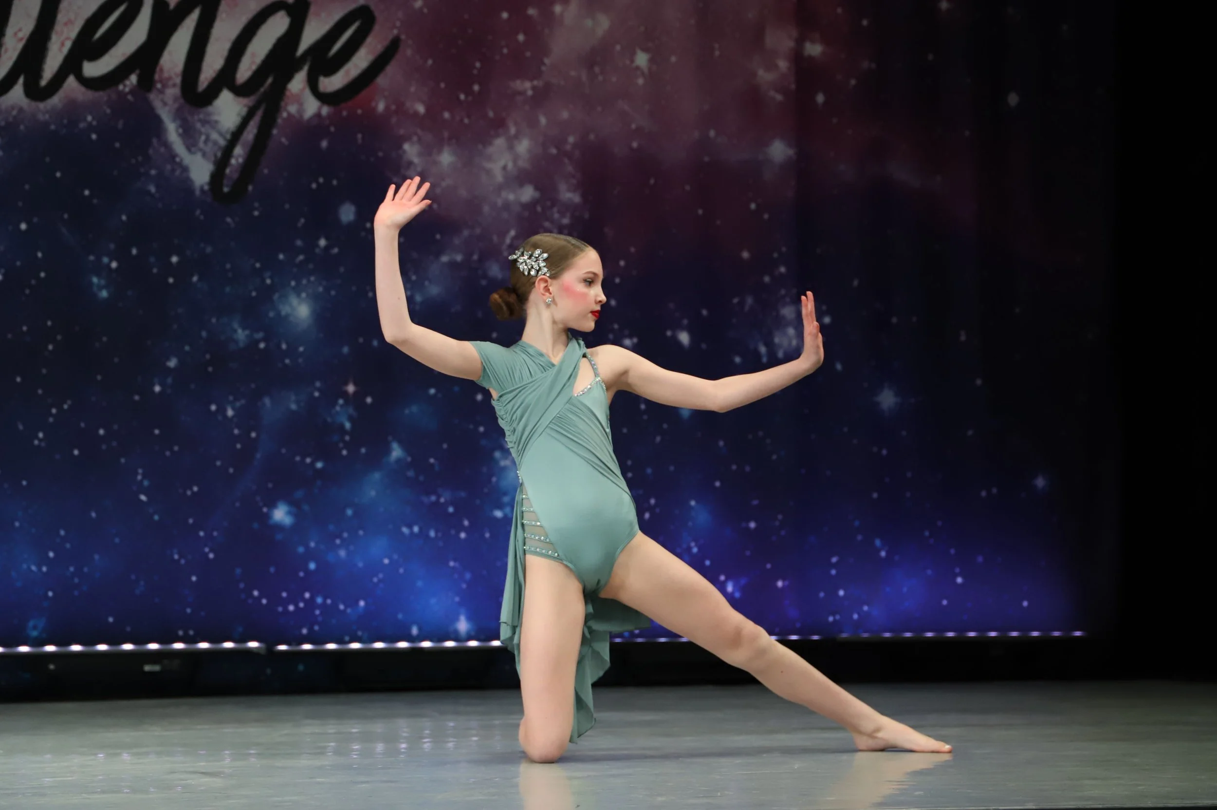A female ballet dancer performing a pose on stage, wearing a light green costume with embellishments, against a starry space-themed backdrop.