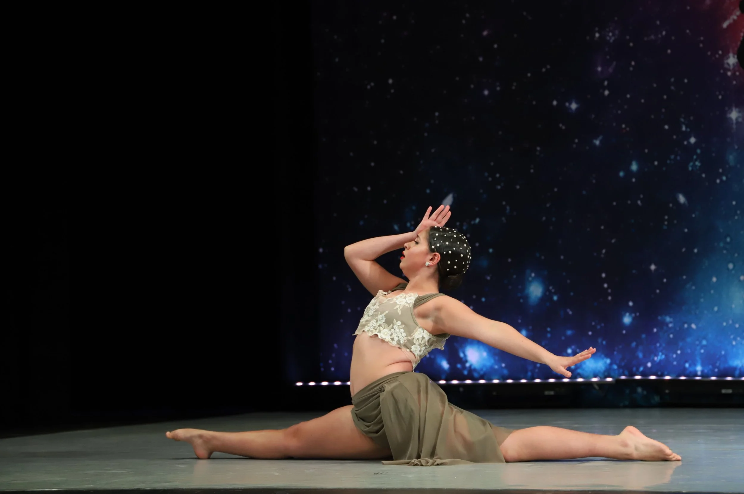 A woman performing a dance pose in front of a starry background, wearing a beige lace top and olive green shorts, with her hair styled in a braided updo decorated with pearls.