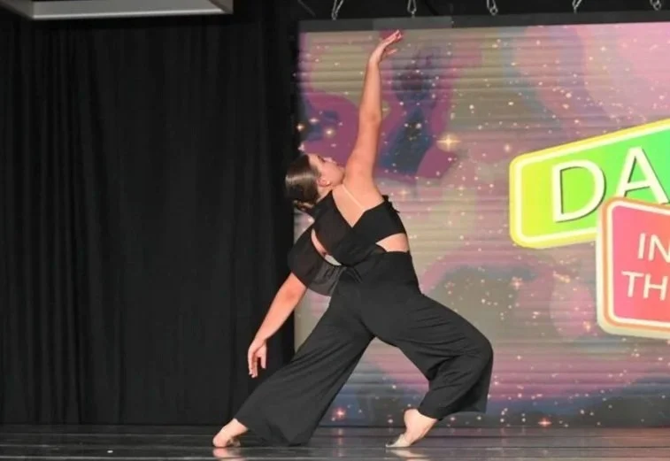 A woman performing a dance move on stage, wearing black crop top and wide-leg pants, with a colorful background and stage curtains.