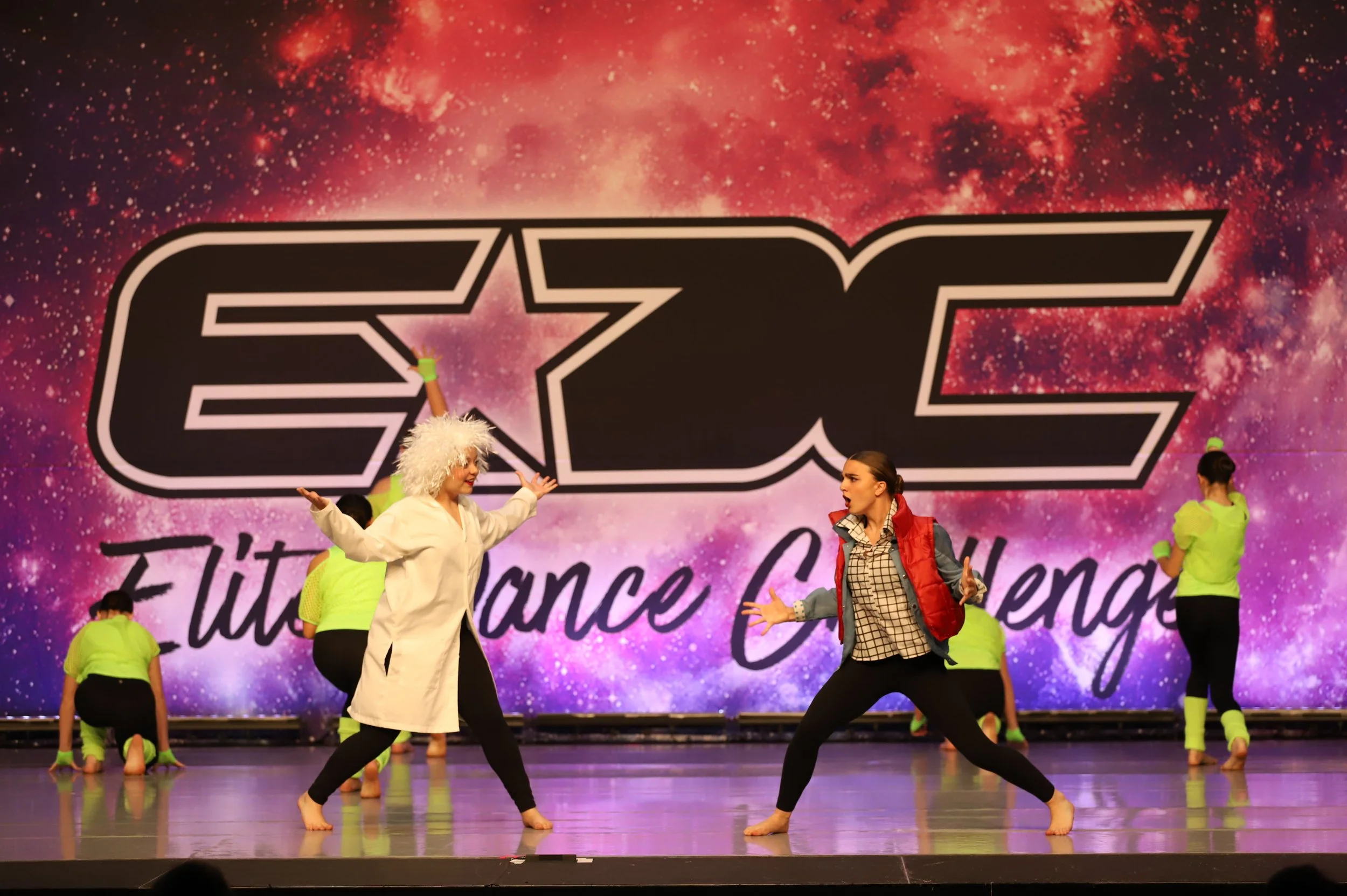 Dance performance on stage with a large backdrop reading 'STC' and 'Title Dance Challenge', featuring two dancers in conversation, one dressed as a scientist with white hair, the other in casual attire, with background dancers in neon yellow outfits