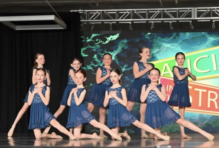 Ten young girls dressed in matching blue dance costumes pose on stage during a performance.