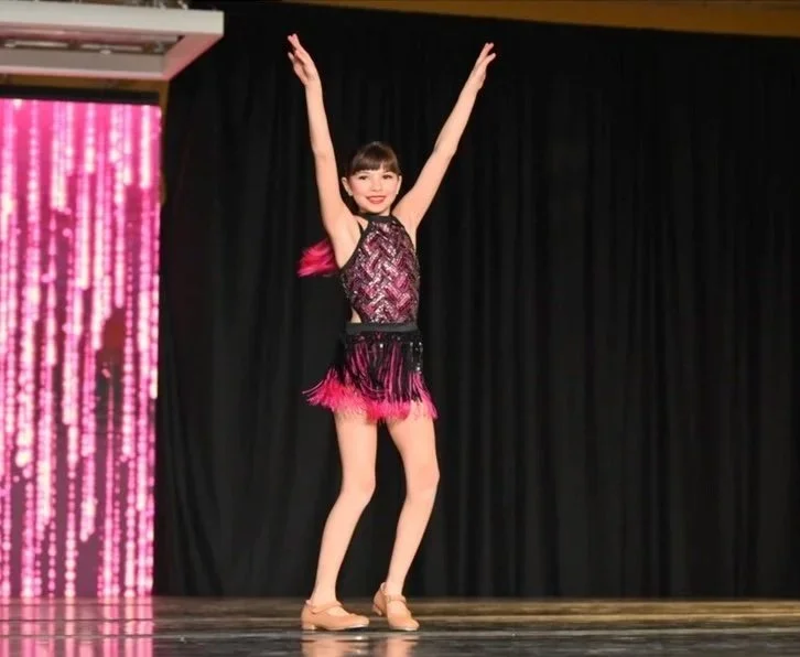 Young girl dancing on stage in a colorful costume with arms raised and a smile, black curtain background.