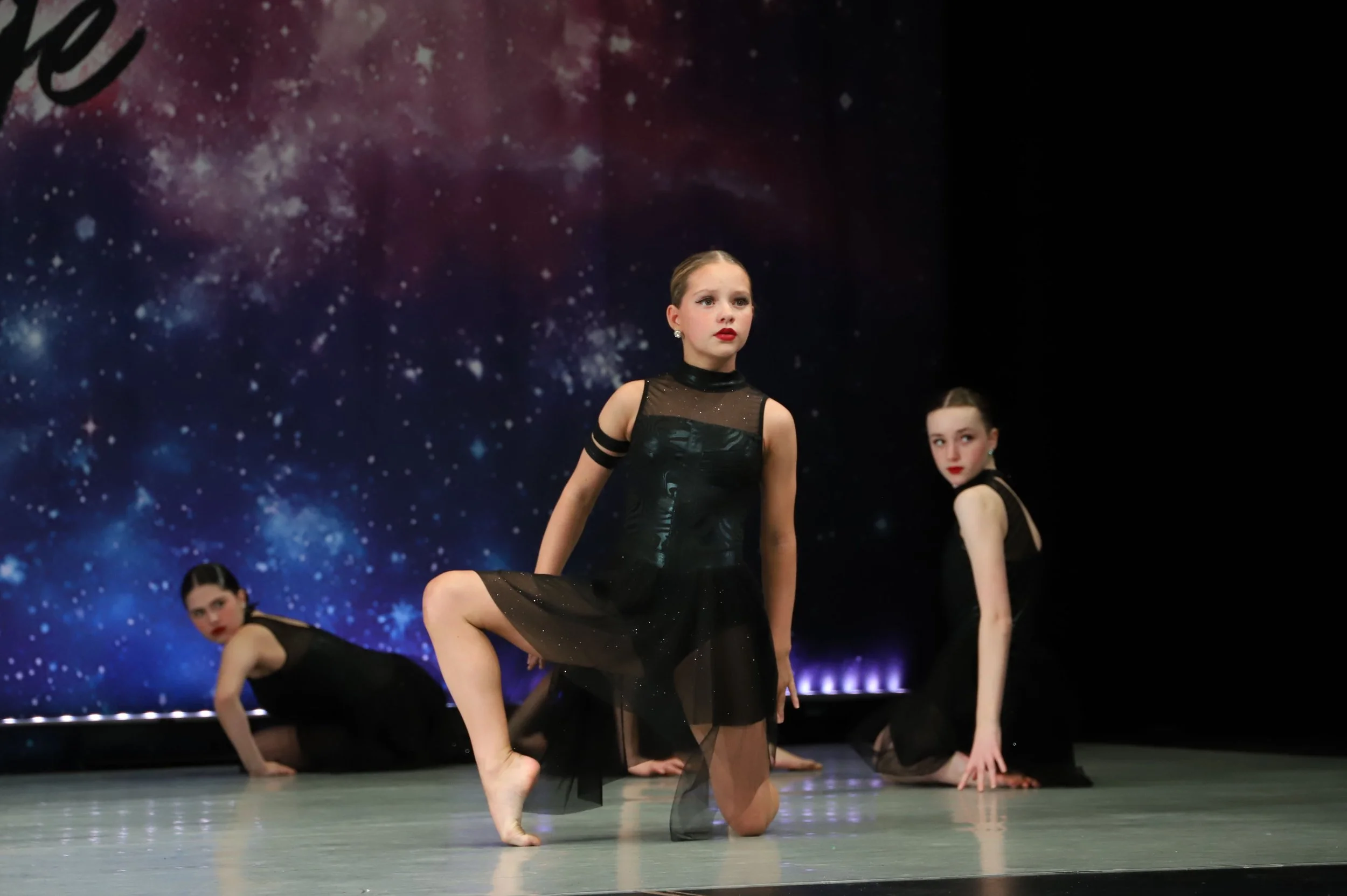 Young girls in black ballet costumes performing a dance routine on stage with a starry galaxy backdrop.