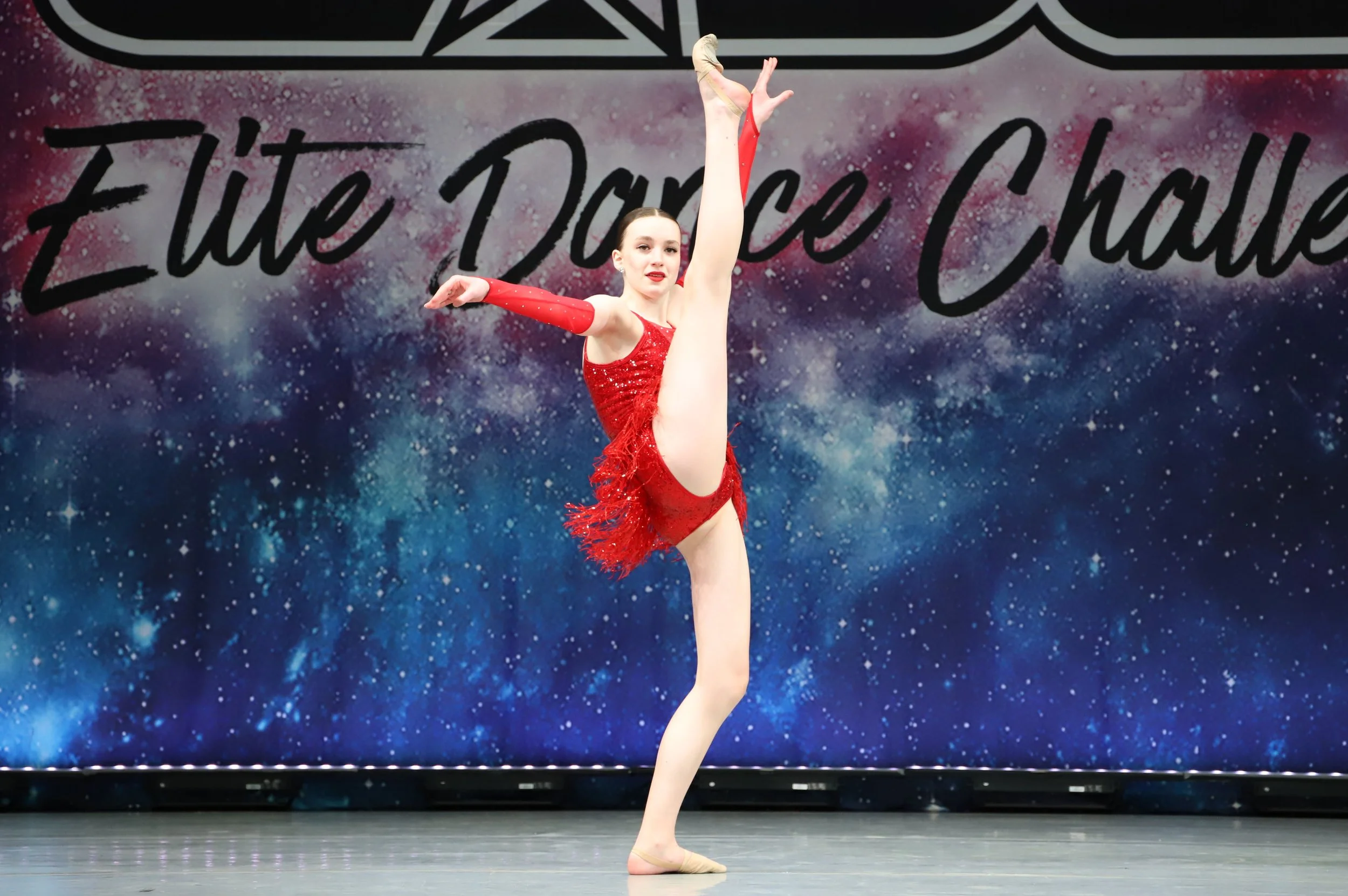A female gymnast in a red leotard performing a balance pose on stage, with a backdrop that reads 'Elite Dance Showcase' and features a cosmic, starry design.