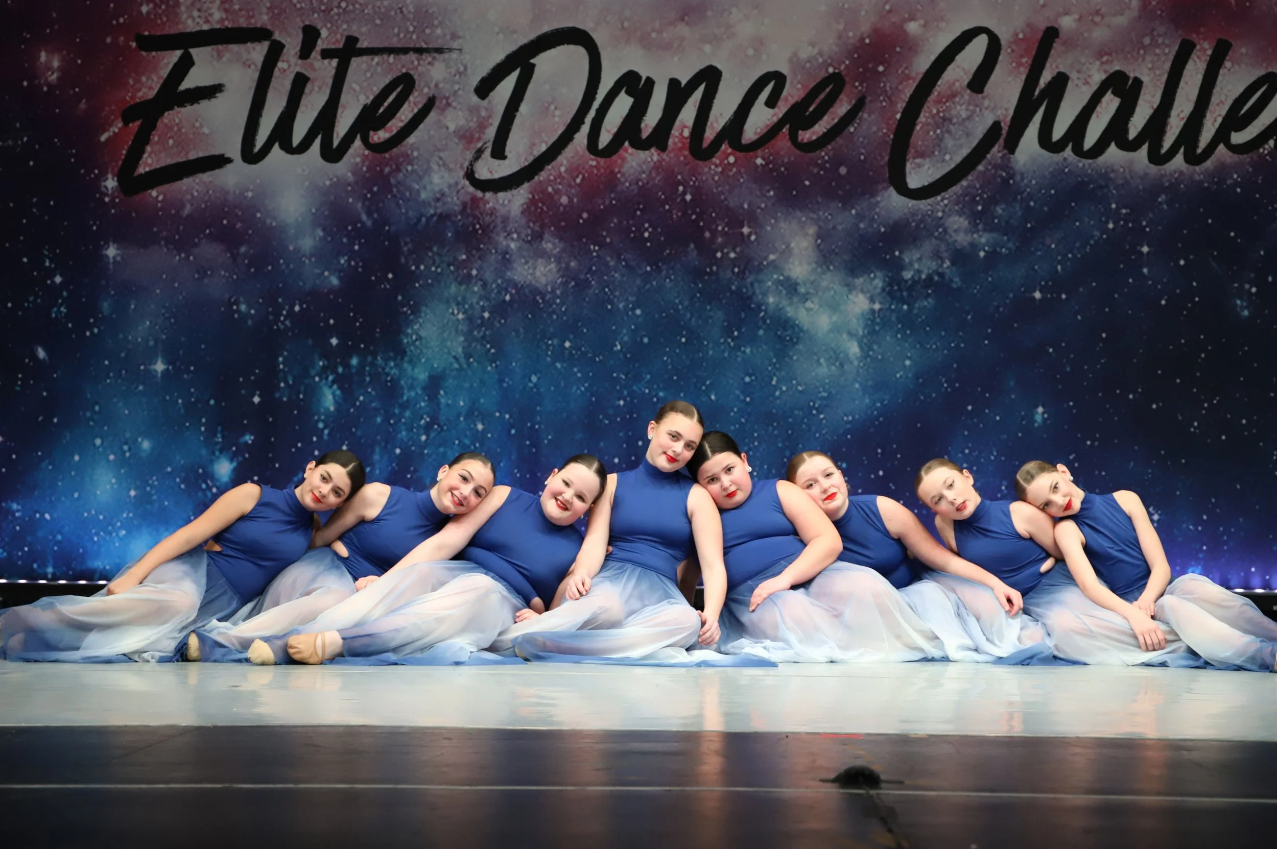Group of young female dancers in matching blue costumes sitting on the stage, leaning on each other, with a starry galaxy backdrop and the words "The Dance I Choose" above.