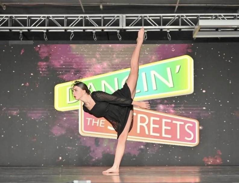 A female dancer in a black dress performing an arabesque on stage with a colorful backdrop reading "DANCIN' IN THE STREETS".