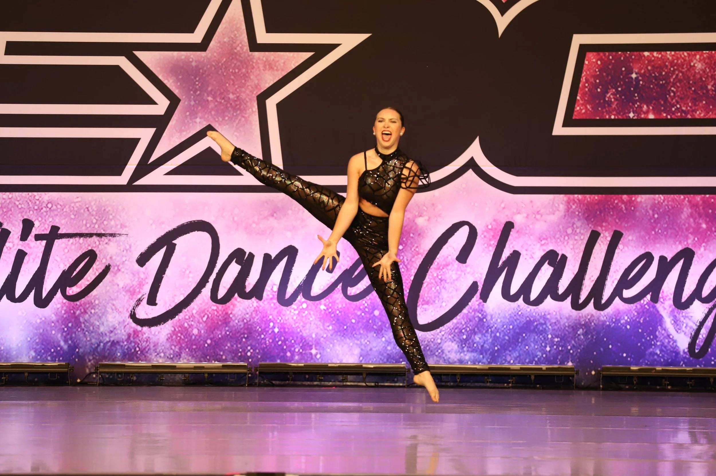 A female dancer performing on stage at the White Dance Challenge, wearing a black, lattice-style dance outfit, with her right leg stretched high and hands touching her right ankle, smiling while looking towards the camera, with a colorful starry back