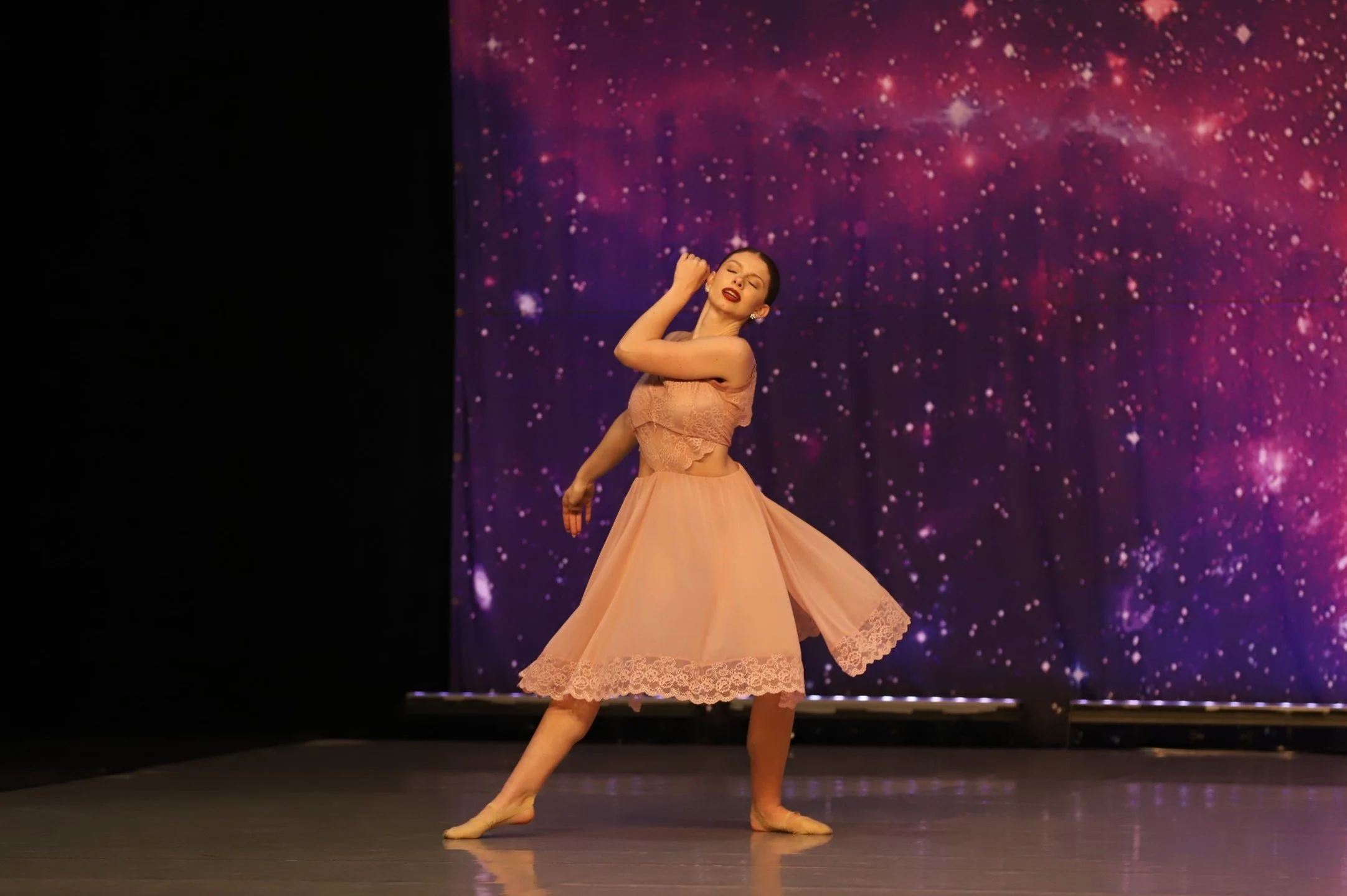A female ballet dancer in a peach dress performing on stage with a cosmic galaxy backdrop.