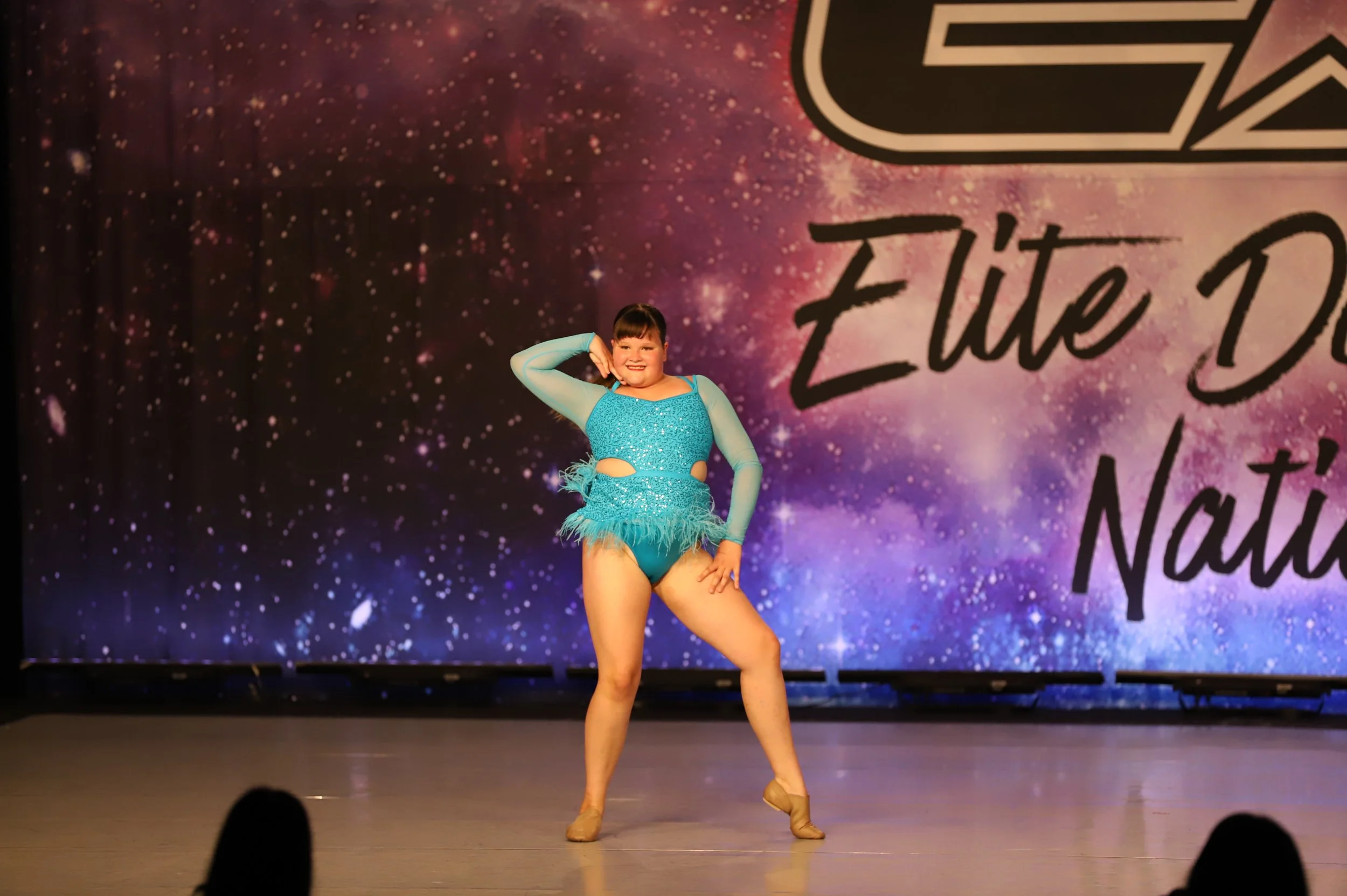 A young girl performing a dance on stage, wearing a bright blue costume with long sleeves and feathered details, standing in front of a space-themed backdrop with text.
