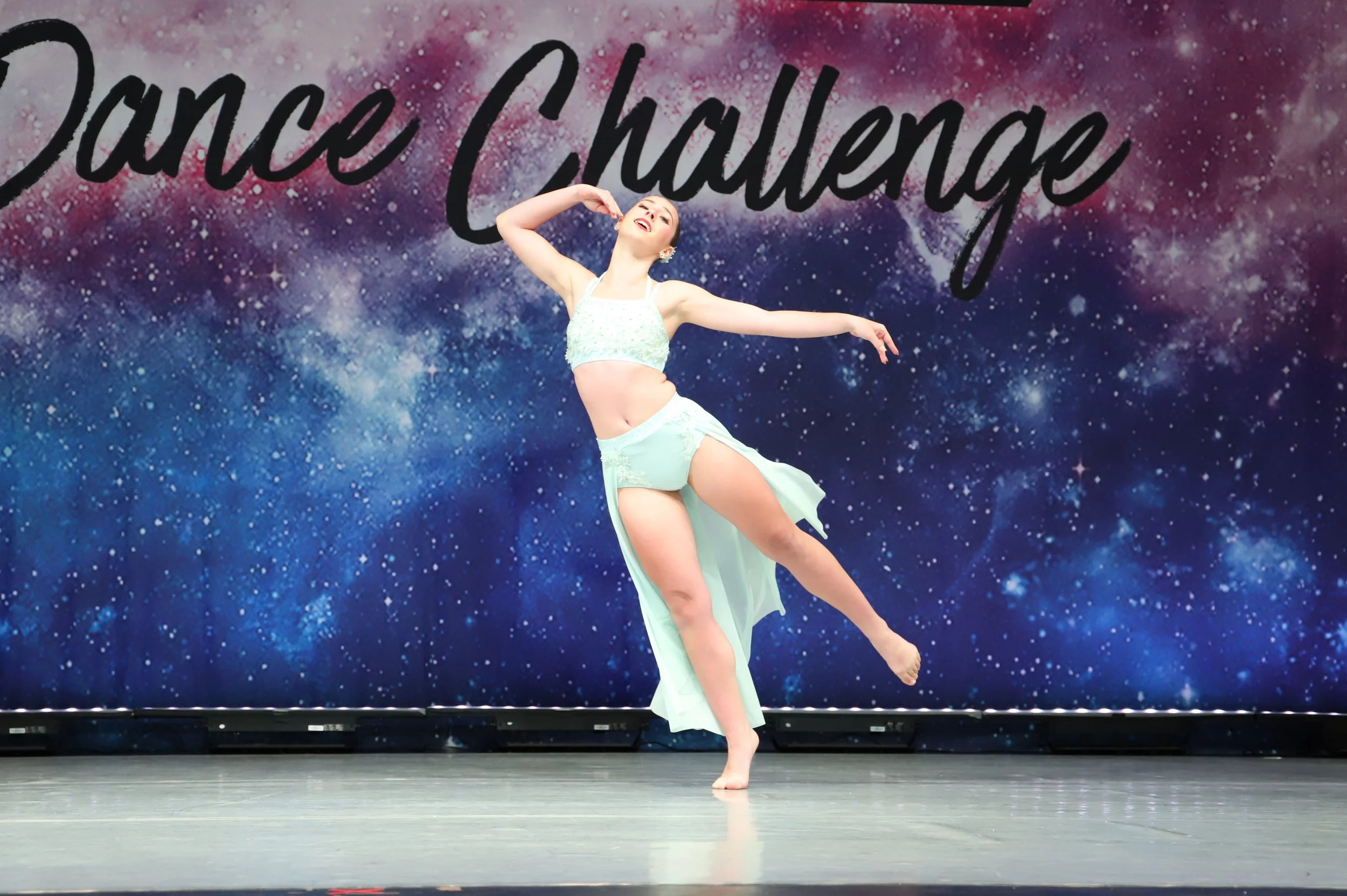 A ballerina performing a dance move on stage with a galaxy-themed backdrop that has the name 'Jace Chourey' written in large cursive letters.