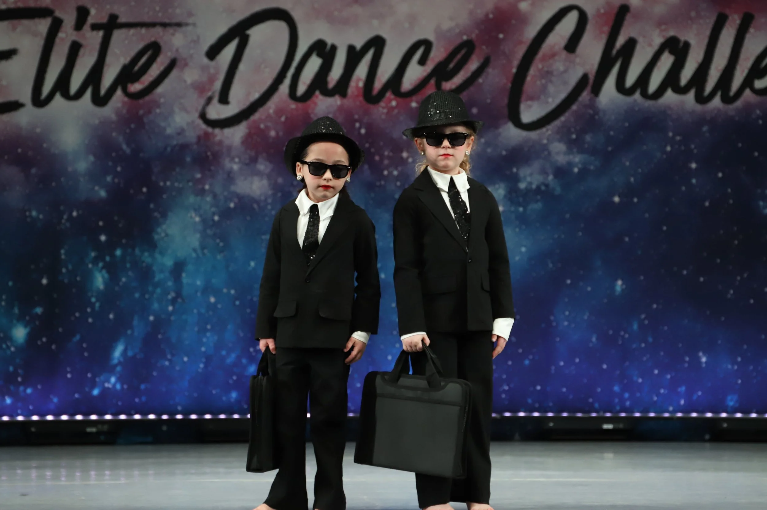 Two young girls dressed in black suits, white shirts, and ties, wearing black hats and sunglasses, standing in front of a colorful galaxy-themed background with the text 'We Dance I Dream'.