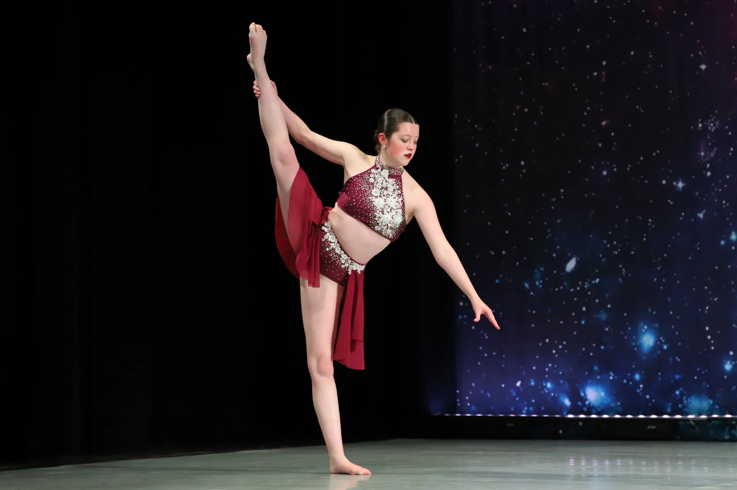 A woman performing a dance pose, standing on one leg, with one arm extended downward and the other arm holding her raised leg behind her head. She is wearing a red costume with white embellishments and a sheer red fabric hanging from her waist, with 