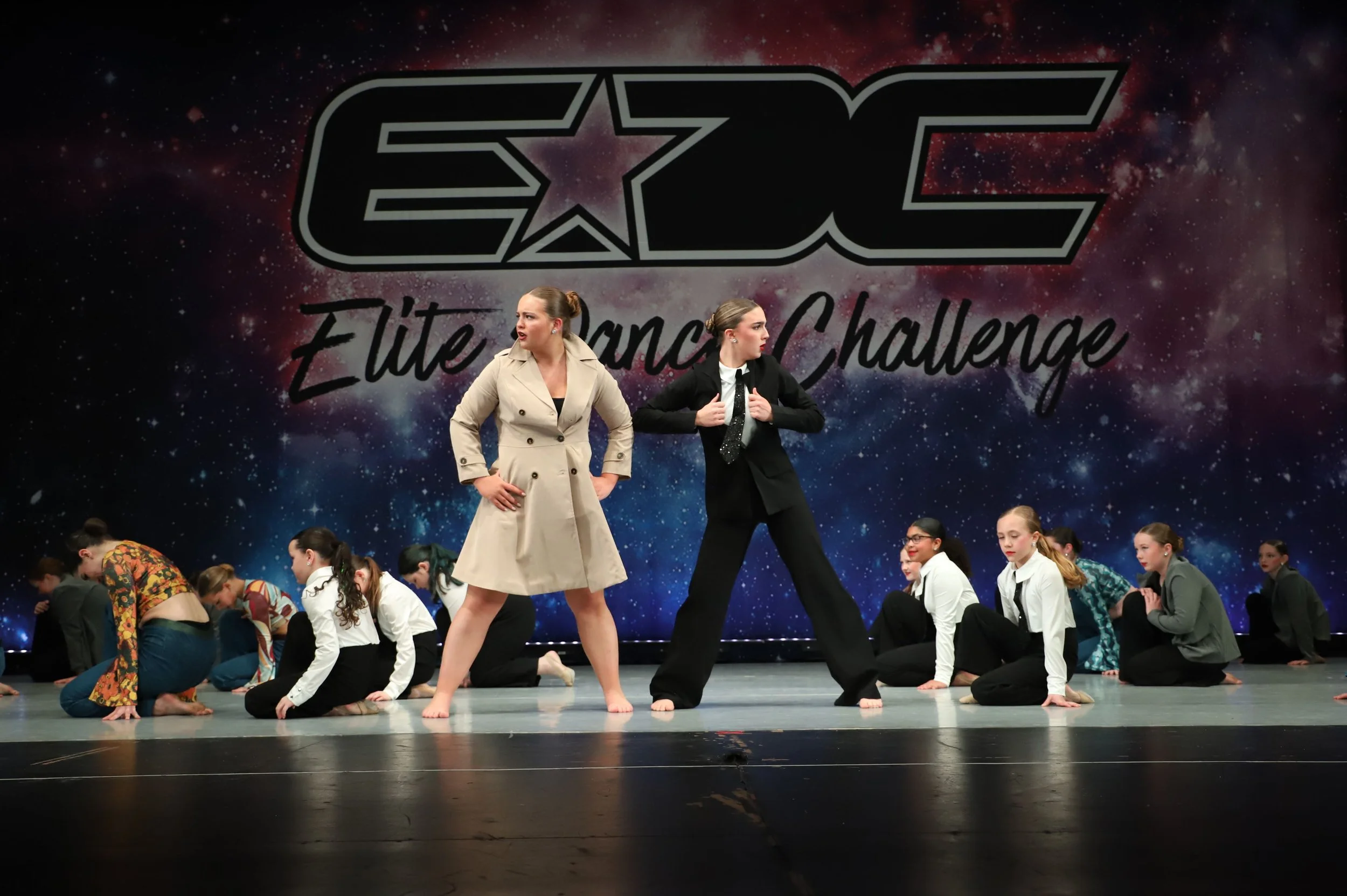Dance performers on stage at a competition with a cosmic-themed backdrop and large logo, with children seated or kneeling on the stage floor.